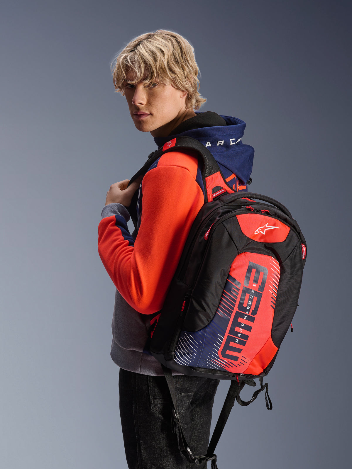 Plecak Alpinestars MM93 City Hunter V2, czarny z czerwonymi fluo i granatowymi akcentami, z grafikami MM93 Marca Marqueza, noszony z pasującą sportową bluzą z kapturem, smukły aerodynamiczny profil do dojazdów motocyklem