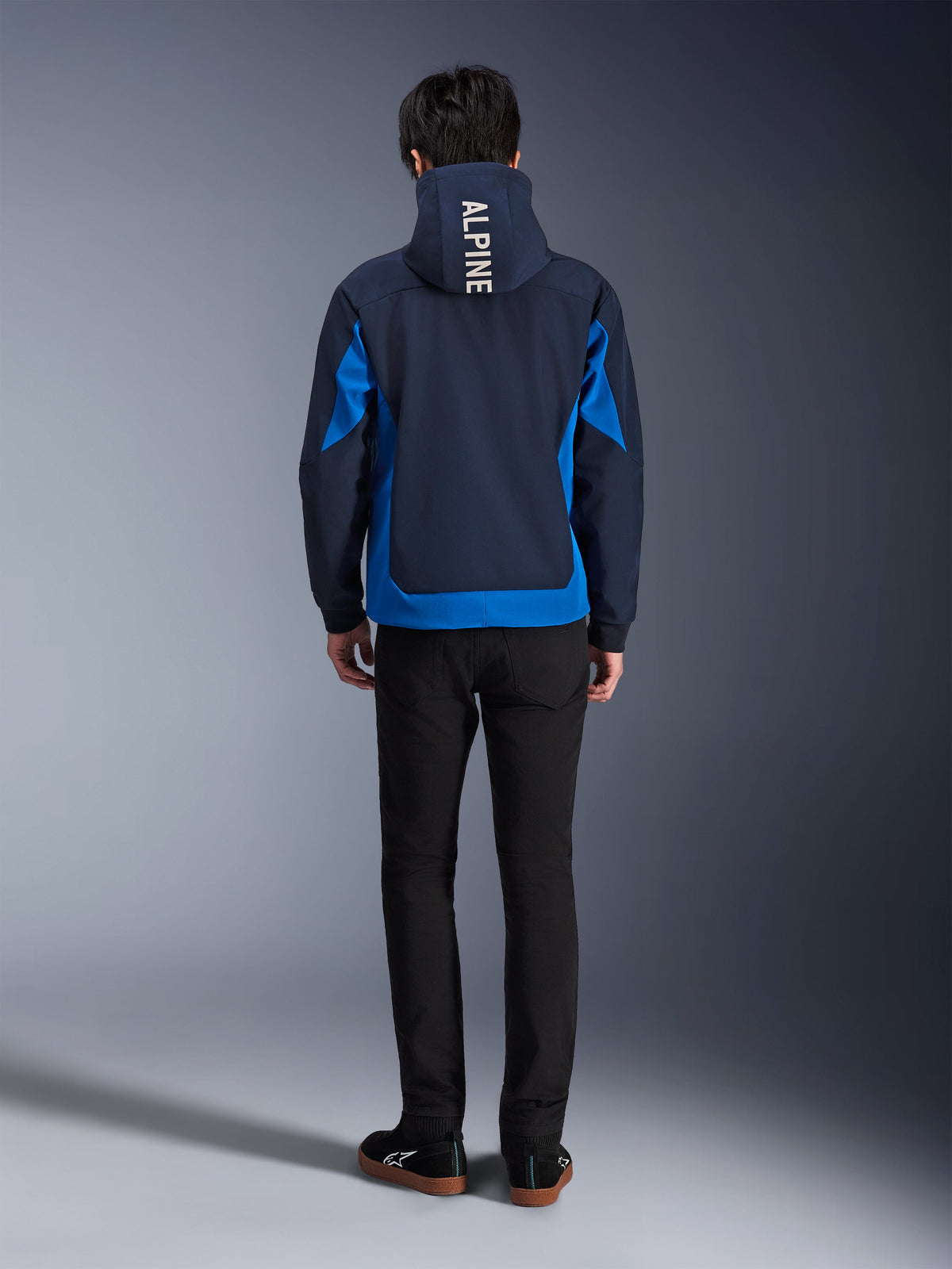 Alpinestars Radium Tech Hoodie, kolor Night Navy i Royal Blue, widok z tyłu z pionowym napisem „ALPINE” na kapturze, niebieskie panele boczne i dolne wykończenie, casualowa miejska bluza motocyklowa z wiatroszczelną powłoką 3L, noszona z czarnymi spodniami
