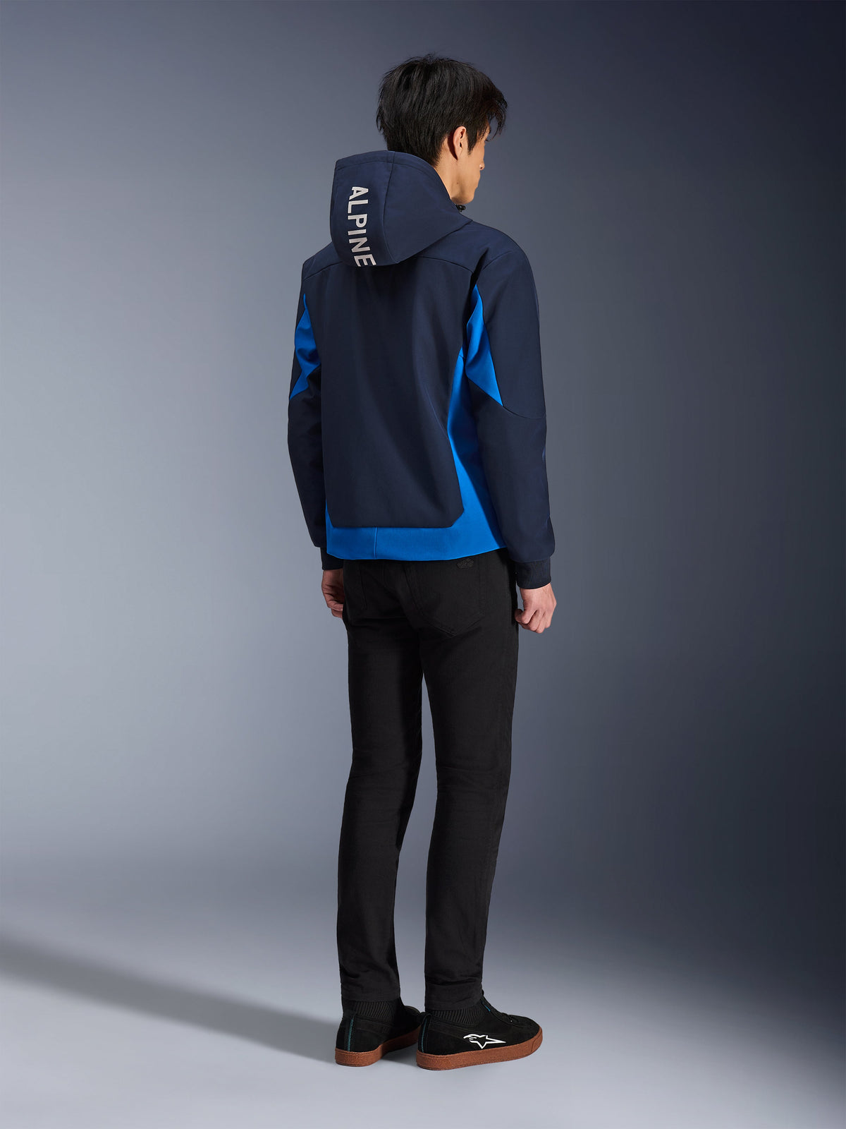 Alpinestars Radium Tech Hoodie, Night Navy i Royal Blue, widok z tyłu modela w technicznej bluzie z kapturem z białym napisem „ALPINE” na kapturze, w zestawie z czarnymi spodniami i butami, miejska odzież motocyklowa
