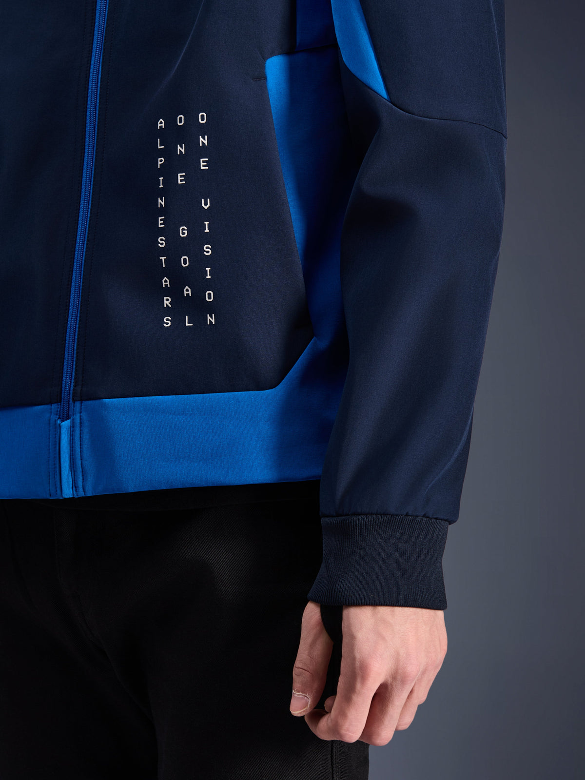 Alpinestars Radium Tech Hoodie, miejska bluza motocyklowa, granatowa i niebieska, zbliżenie na panel boczny z białą pionową grafiką tekstową, elastyczny prążkowany mankiet i niebieskie zapięcie na zamek
