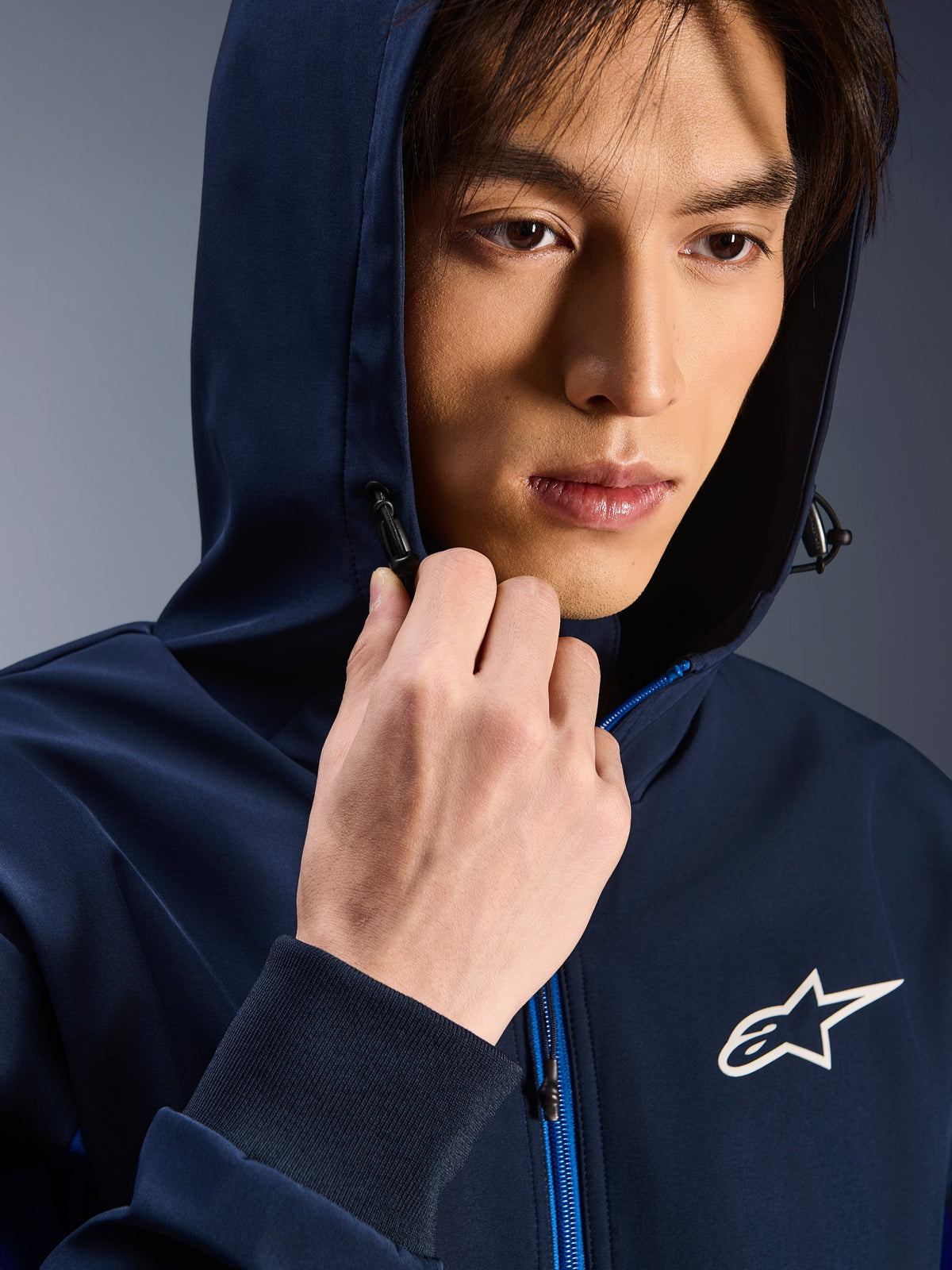 Alpinestars Radium Tech Hoodie, Night Navy Royal Blue, zbliżenie na mężczyznę w bluzie z założonym kapturem, białe logo Astars na piersi, niebieski zamek błyskawiczny, miejska techniczna odzież motocyklowa