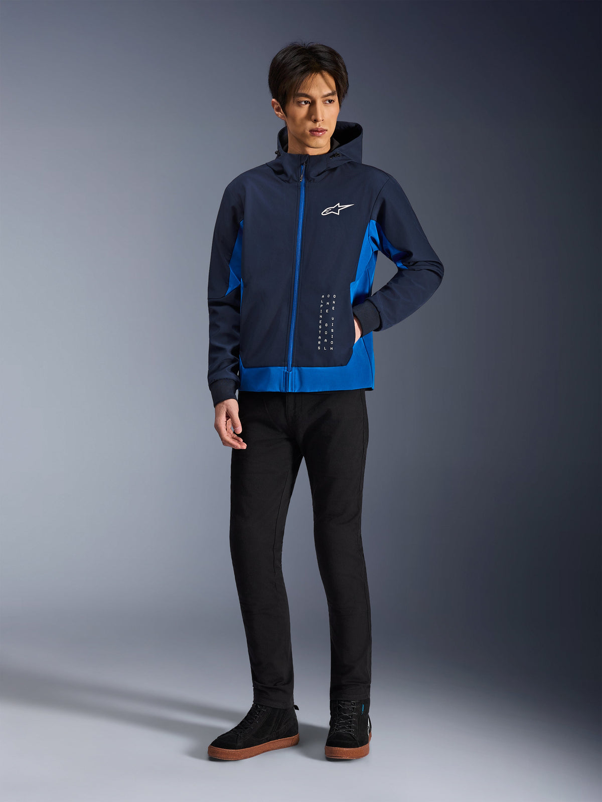 Alpinestars Radium Tech Hoodie, miejska kurtka motocyklowa, granatowa i niebieska (Night Navy i Royal Blue), noszona z czarnymi spodniami i czarnymi sneakersami, białe logo Alpinestars na klatce piersiowej i pionowe napisy, casualowy sportowy krój do jazdy miejskiej