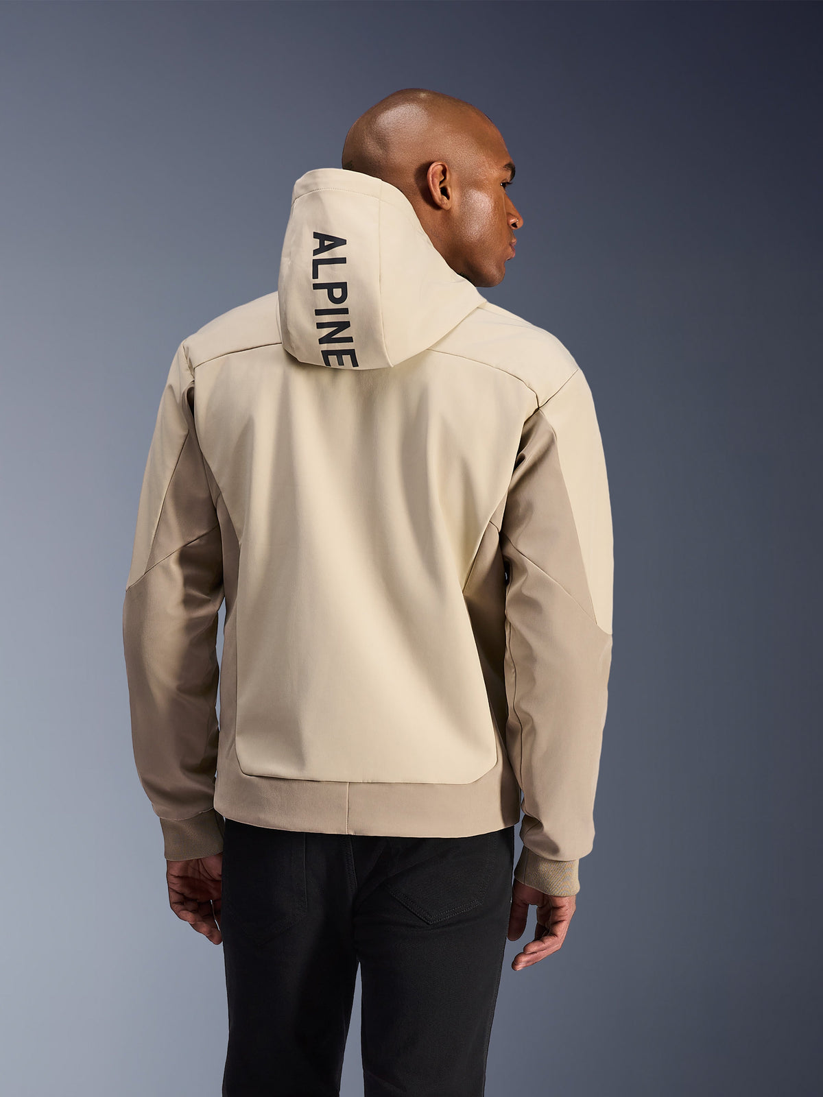 Alpinestars Radium Tech Hoodie, rear view of a man wearing the Mastic Aluminum technical hoodie, featuring vertical ALPINE text on the hood, light beige and tan paneling, ribbed cuffs, urban motorcycle riding jacket