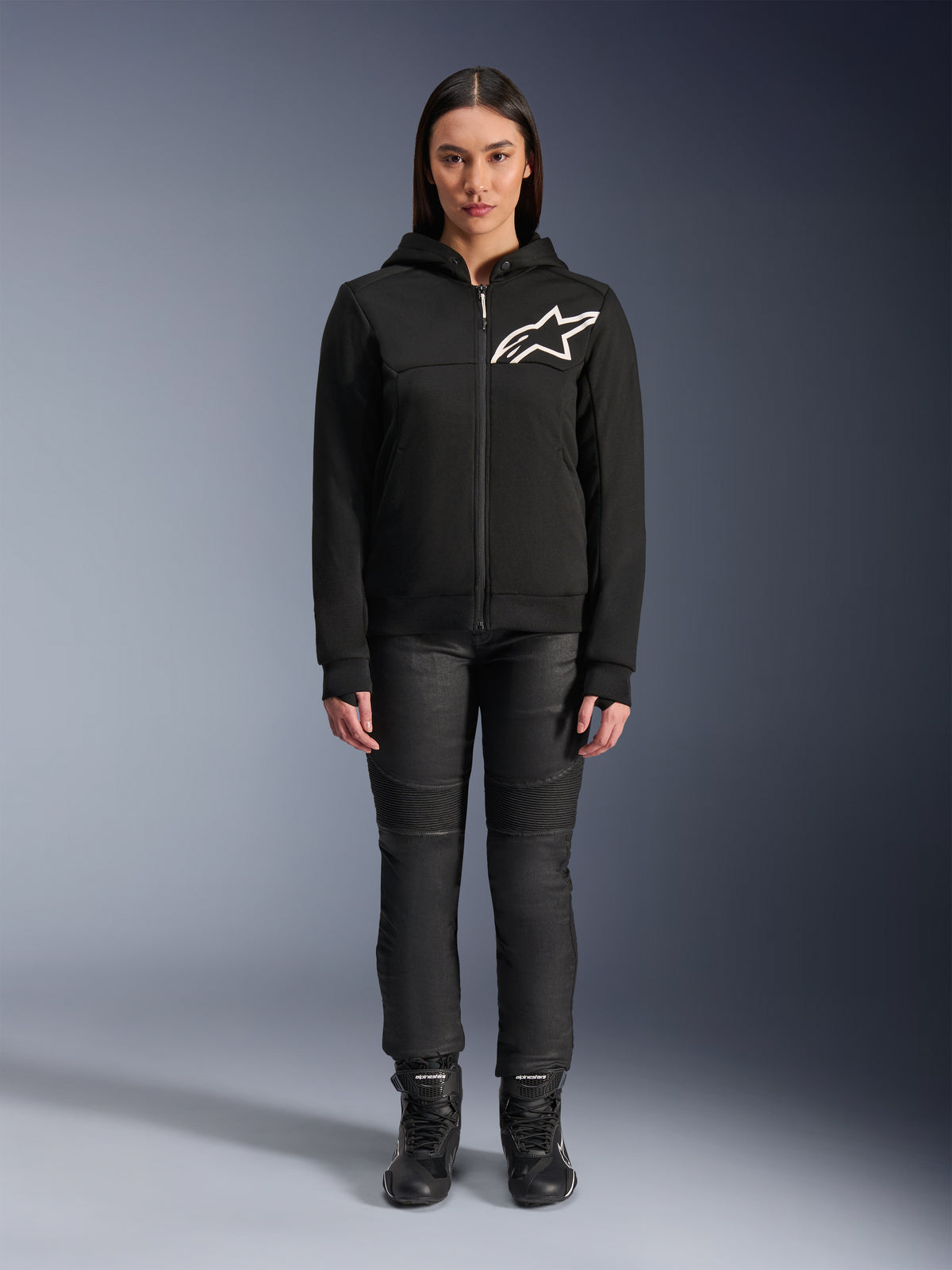 Kobieta w bluzie z kapturem Alpinestars Stella Chrome V2 Sport Hoodie, czarna z białym logo Astars na klatce piersiowej, zestawiona z czarnymi technicznymi spodniami motocyklowymi i butami, miejski styl odzieży motocyklowej.