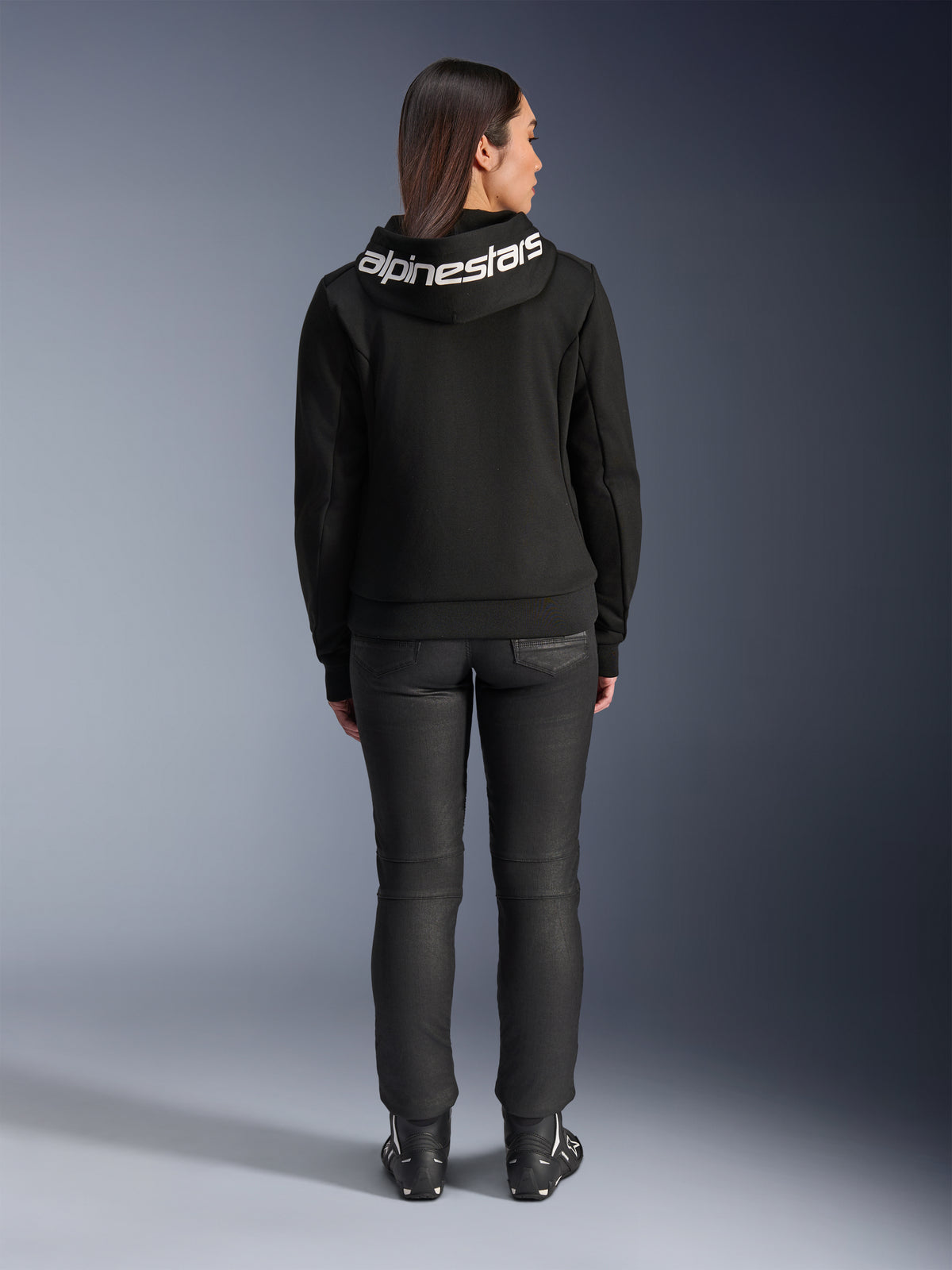 Widok z tyłu modelki ubranej w czarną bluzę Alpinestars Stella Chrome V2 Sport Hoodie, z wyraźnym białym logo Alpinestars na kapturze, zestawionej z czarnymi spodniami do jazdy i butami motocyklowymi, miejska sportowa odzież motocyklowa dla kobiet