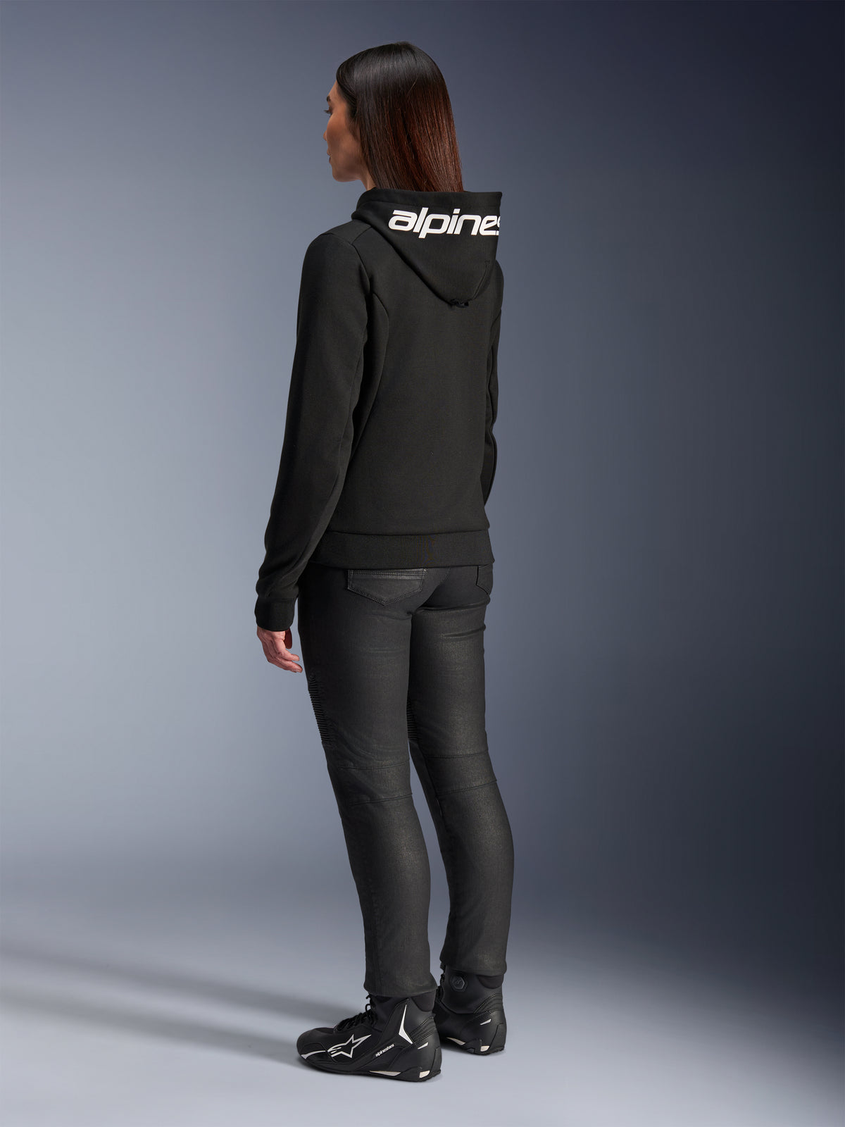 Kobieta ubrana w bluzę Alpinestars Stella Chrome V2 Sport Hoodie, widok z tyłu, czarna motocyklowa bluza z kapturem z białym logo Alpinestars na kapturze, zestawiona z czarnymi technicznymi spodniami do jazdy i czarnymi butami motocyklowymi, casualowy miejski strój do dojazdów