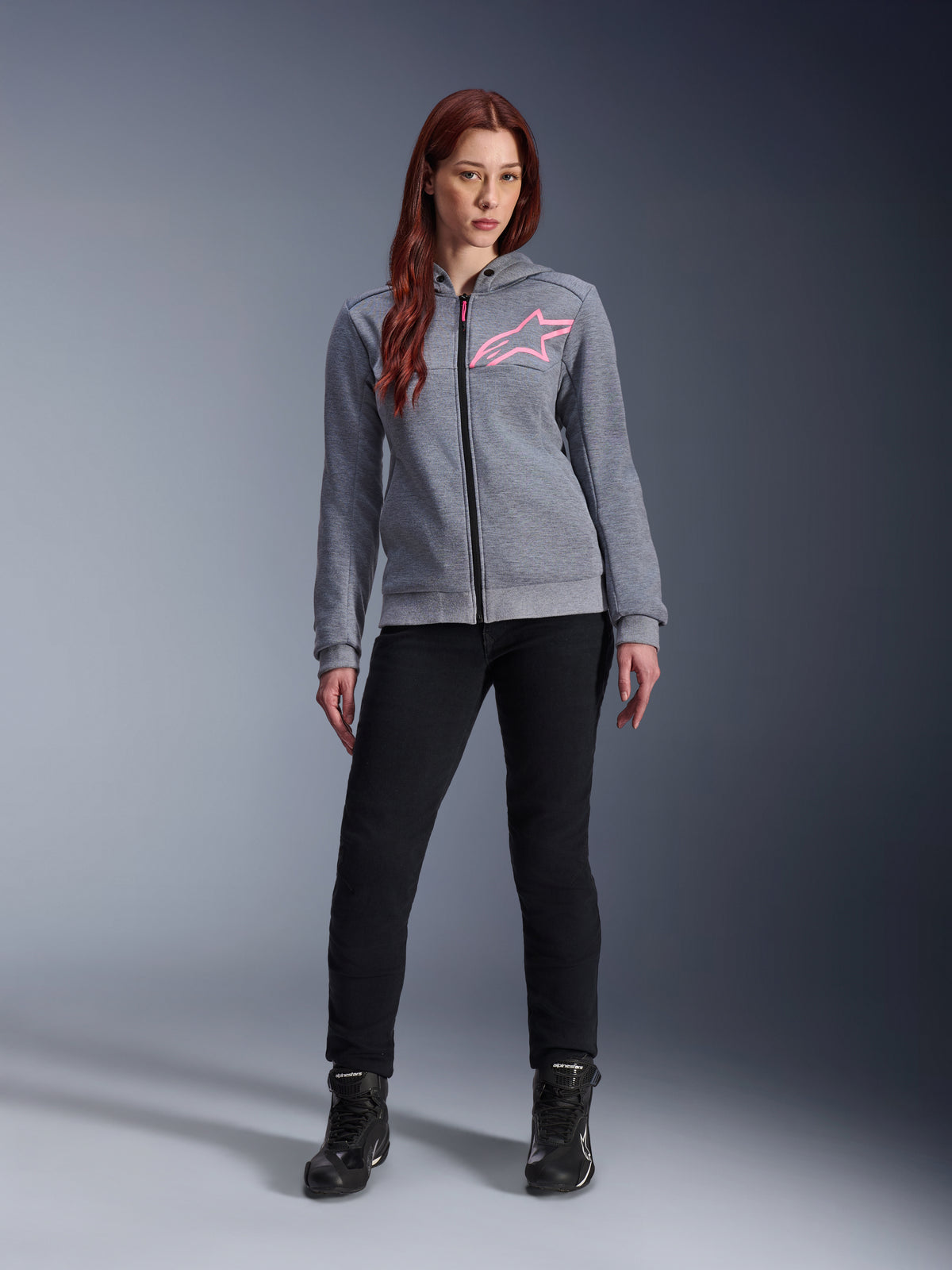 Alpinestars Stella Chrome V2 Sport Hoodie, kolor szary melanż z neonowym różem, noszona przez kobietę w czarnych spodniach i butach, różowe logo gwiazdy Alpinestars na piersi, miejska odzież motocyklowa z pełnym zamkiem, lekki polar techniczny o sportowym kroju.