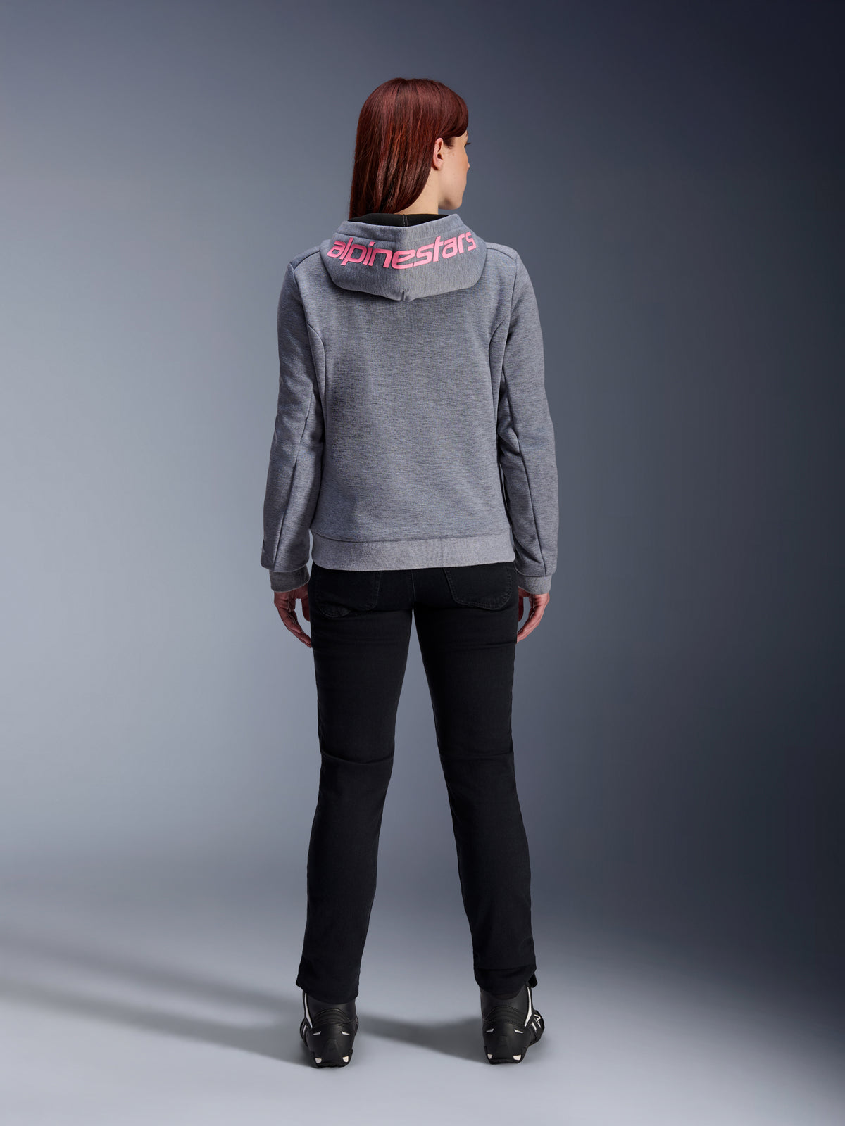Alpinestars Stella Chrome V2 Sport Hoodie, damska bluza motocyklowa, kolor szary melanż i różowy fluo, widok z tyłu prezentujący różowe logo Alpinestars na kapturze, noszona z czarnymi spodniami i butami motocyklowymi, codzienna odzież motocyklowa dla kobiet