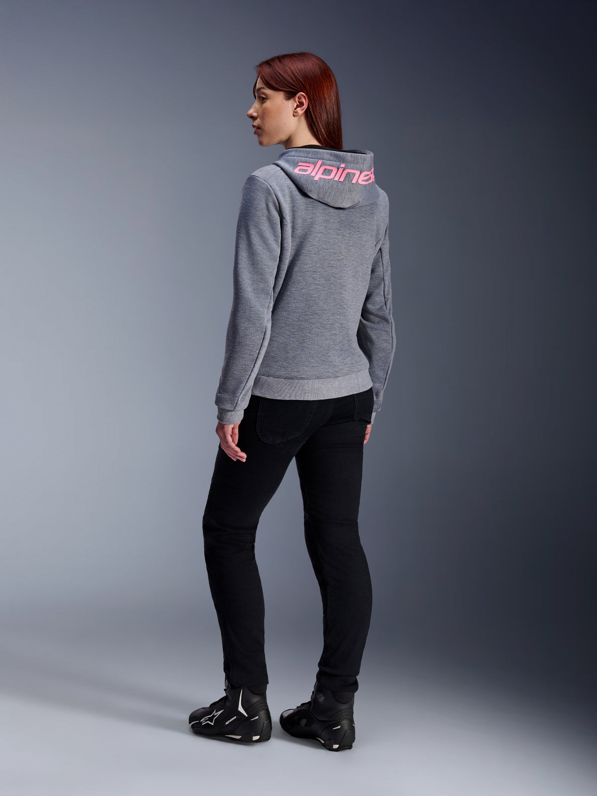 Alpinestars Stella Chrome V2 Sport Hoodie, damska bluza motocyklowa, szary melanż z różowym neonowym brandingiem na kapturze, widok z tyłu, zestawiona z czarnymi spodniami i butami motocyklowymi, swobodny miejski styl z ochroną motocyklową