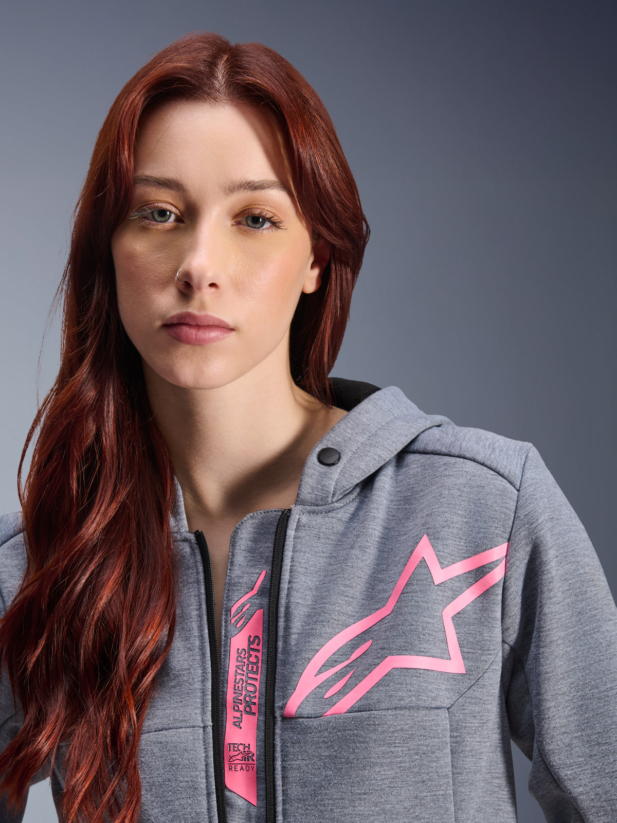 Kobieta w bluzie Alpinestars Stella Chrome V2 Sport Hoodie, Gray Heather Pink Fluo, zbliżenie na różowe logo gwiazdy Alpinestars oraz oznaczenie Tech-Air Ready, miejska odzież motocyklowa