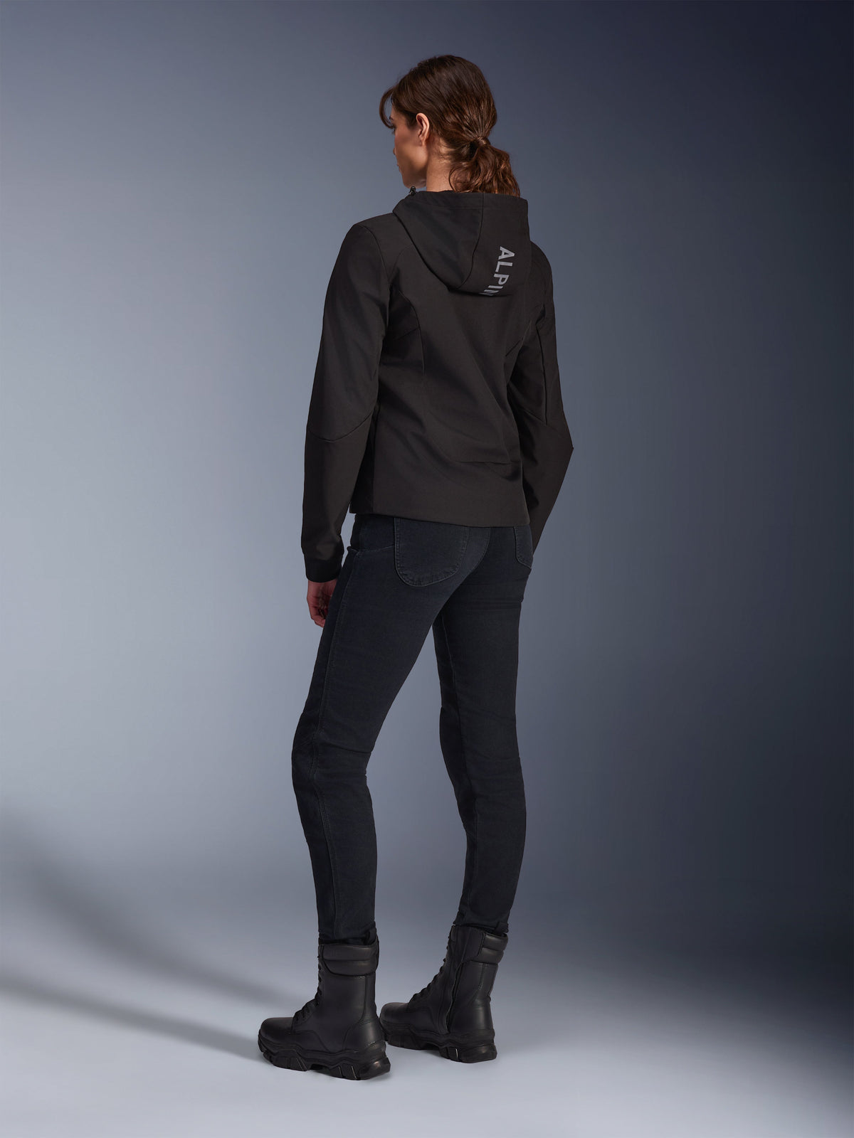 Alpinestars Stella Radium Tech Hoodie, czarna damska bluza motocyklowa do jazdy miejskiej, widok z tyłu prezentujący napis „ALPIN” na kapturze, zestawiona z czarnymi technicznymi spodniami motocyklowymi i czarnymi butami, lekka wiatroszczelna powłoka techniczna.