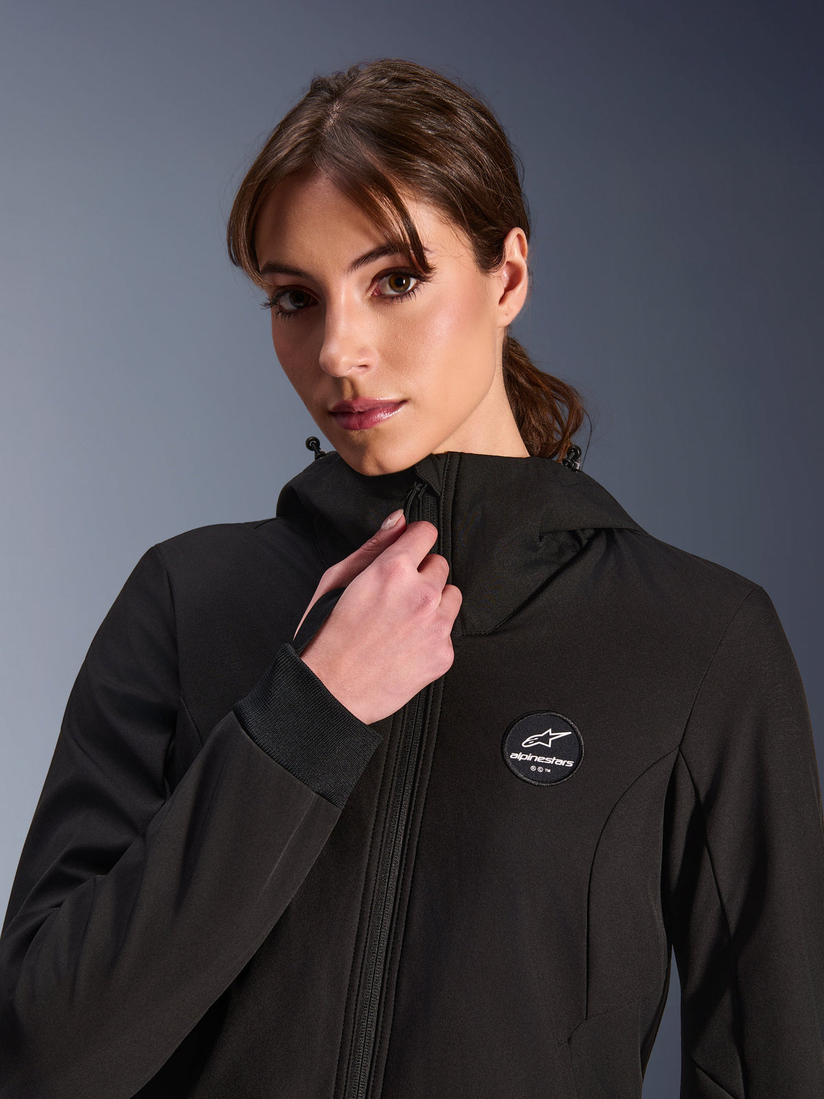 Damska bluza Stella Radium Tech Hoodie, czarna, noszona przez kobietę regulującą przedni zamek, z okrągłą naszywką z logo Alpinestars na klatce piersiowej, żebrowanymi elastycznymi mankietami i wiatroszczelną konstrukcją tekstylną do jazdy miejskiej