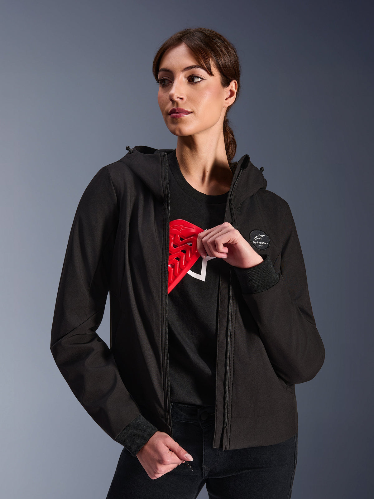 Alpinestars Woman Stella Radium Tech Hoodie, czarna miejska bluza motocyklowa z kapturem, modelka prezentująca czerwony ochraniacz Nucleon Flex Plus wewnątrz, lekka 3-warstwowa wiatroszczelna powłoka tekstylna ze ściągaczami i naszywką z logo na piersi, ochronna odzież motocyklowa