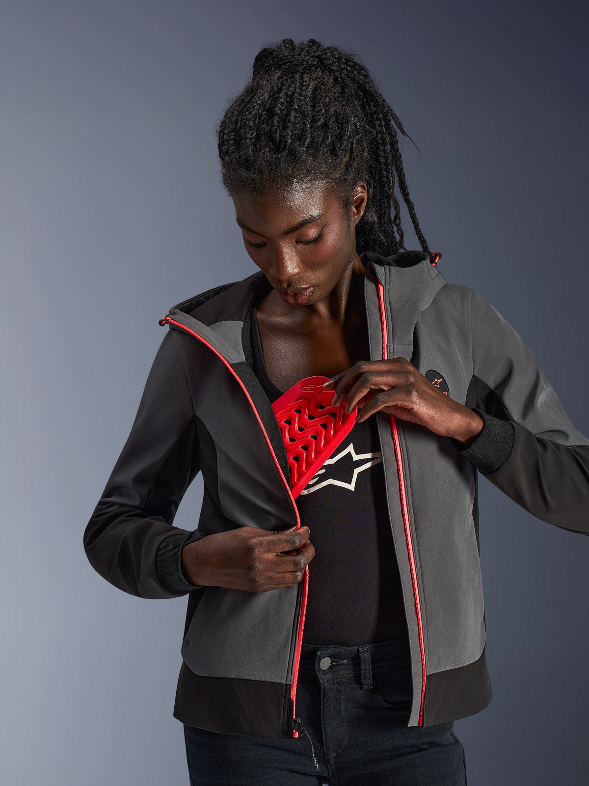 Alpinestars Stella Radium Tech Hoodie, damska bluza motocyklowa w kolorze ciemnoszarym i czarnym, modelka prezentująca wyjmowany czerwony ochraniacz Nucleon Flex Plus, miejska bluza z różowymi akcentami przy zamkach