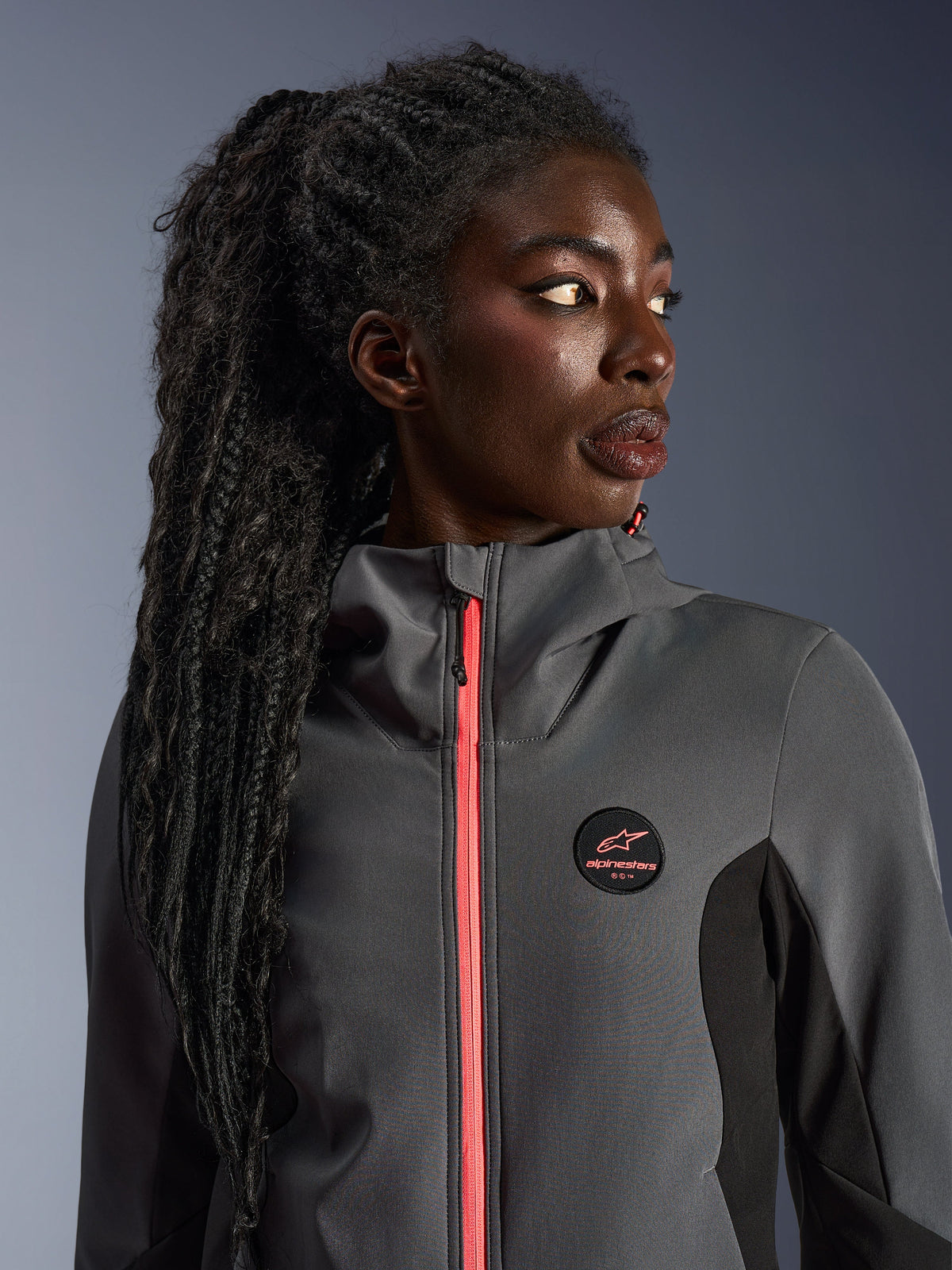 Modelka w bluzie Alpinestars Stella Radium Tech Hoodie w kolorze ciemnoszarym i czarnym, z jaskrawym koralowym zamkiem, okrągłym logo na piersi i wiatroszczelną powłoką z wysokim kołnierzem, miejska odzież motocyklowa dla kobiet