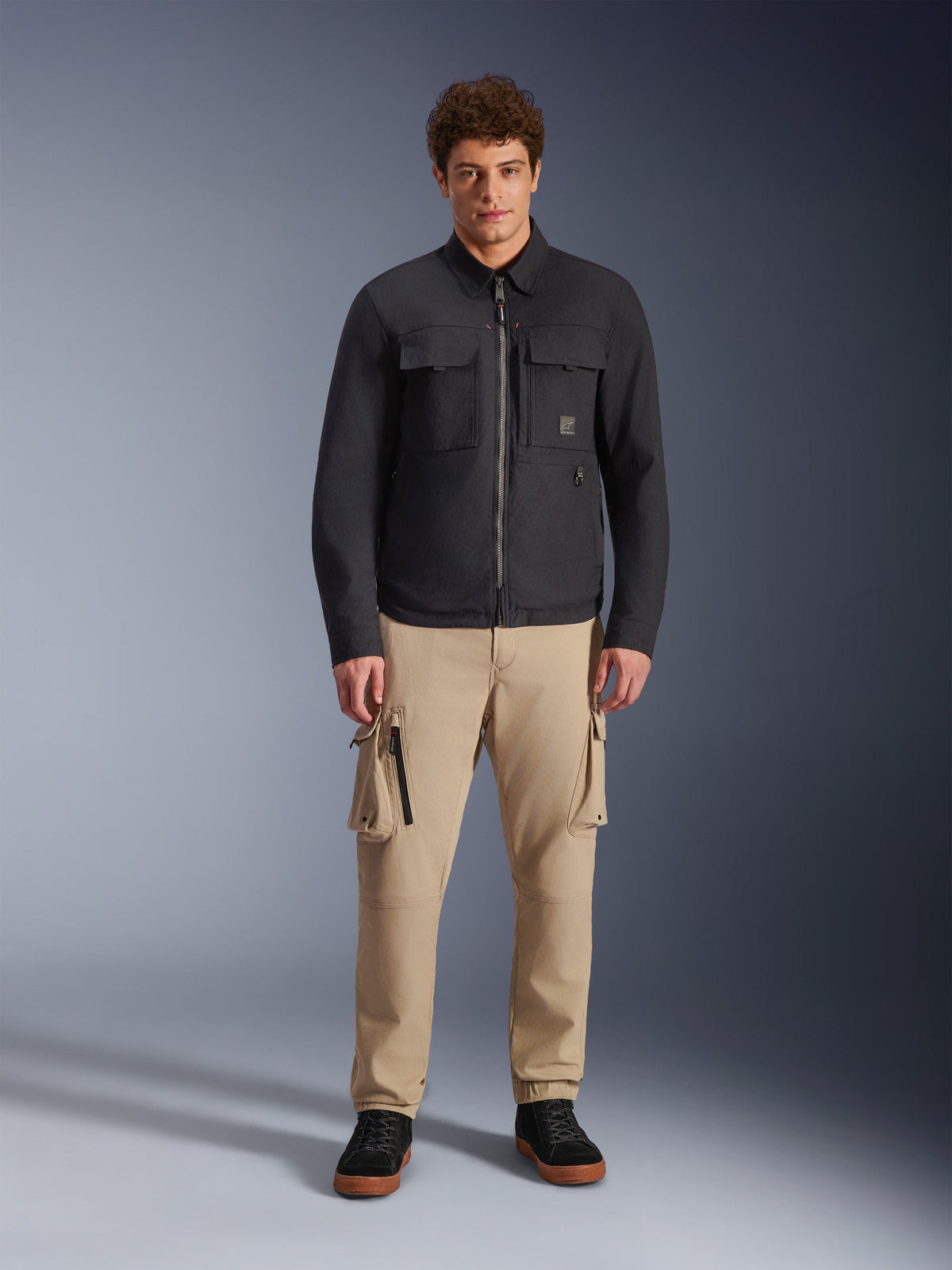 Alpinestars Bryon Water Resistant Overshirt, czarna męska kurtka koszulowa z kieszeniami cargo i centralnym zamkiem, beżowe spodnie cargo, czarne sneakersy, portret studyjny całej sylwetki.