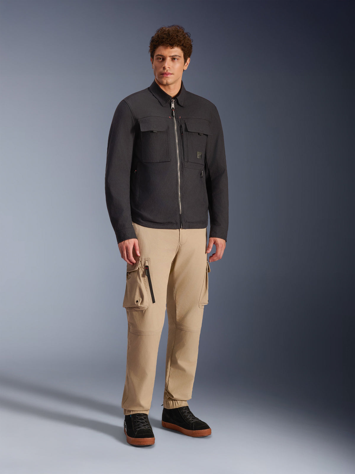Alpinestars Bryon Water Resistant Overshirt, czarna, prezentowana przez modela w spodniach cargo khaki i czarnych butach, miejska kurtka motocyklowa o designie inspirowanym odzieżą roboczą, ujęcie studyjne całej sylwetki