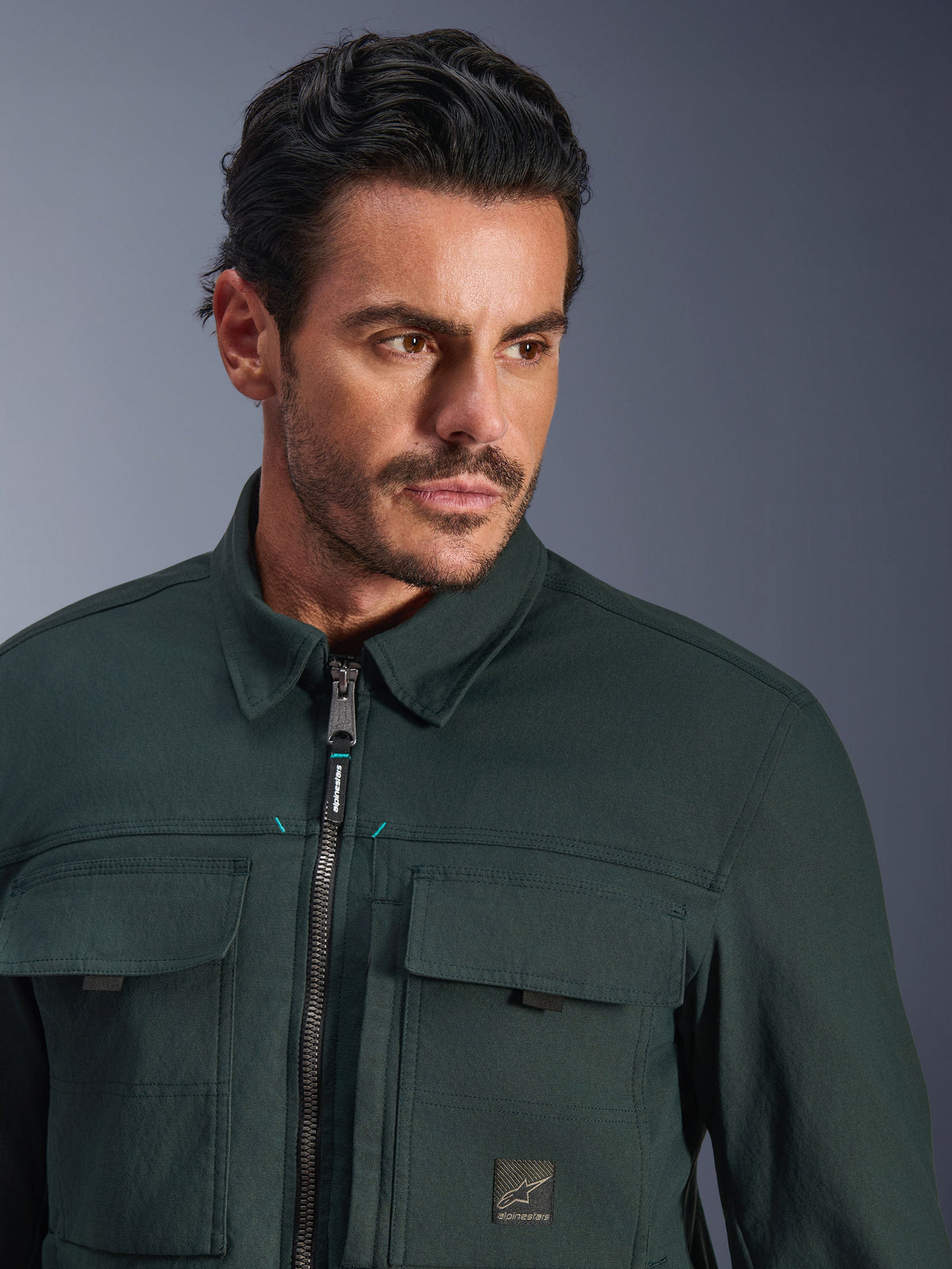 Alpinestars Bryon Water Resistant Overshirt in Dark Emerald, close-up portrait of a man wearing the urban motorcycle jacket, featuring waxed canvas texture, front zipper with subtle teal accents, chest cargo pockets, and branded logo patch.