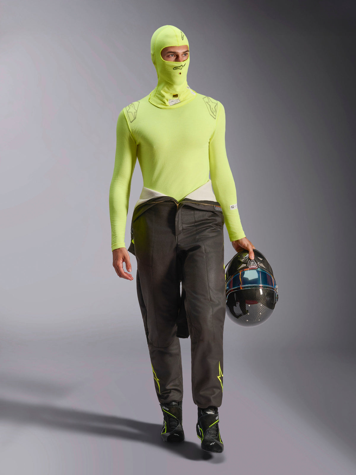 Male racing driver wearing Alpinestars ZX Evo V3 Balaclava in Yellow Fluo, matching long-sleeve base layer top, dark racing pants, and black racing boots, holding a carbon fiber helmet, professional automotive racing gear