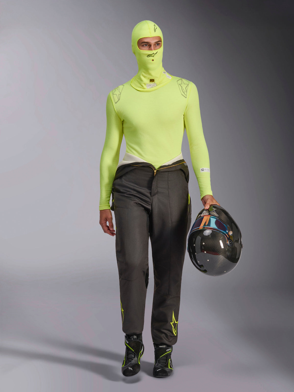 Alpinestars ZX Evo V3 Balaclava in Yellow Fluo, worn with matching long-sleeve base layer top and dark grey racing pants, male model holding a carbon fiber helmet, professional auto racing gear