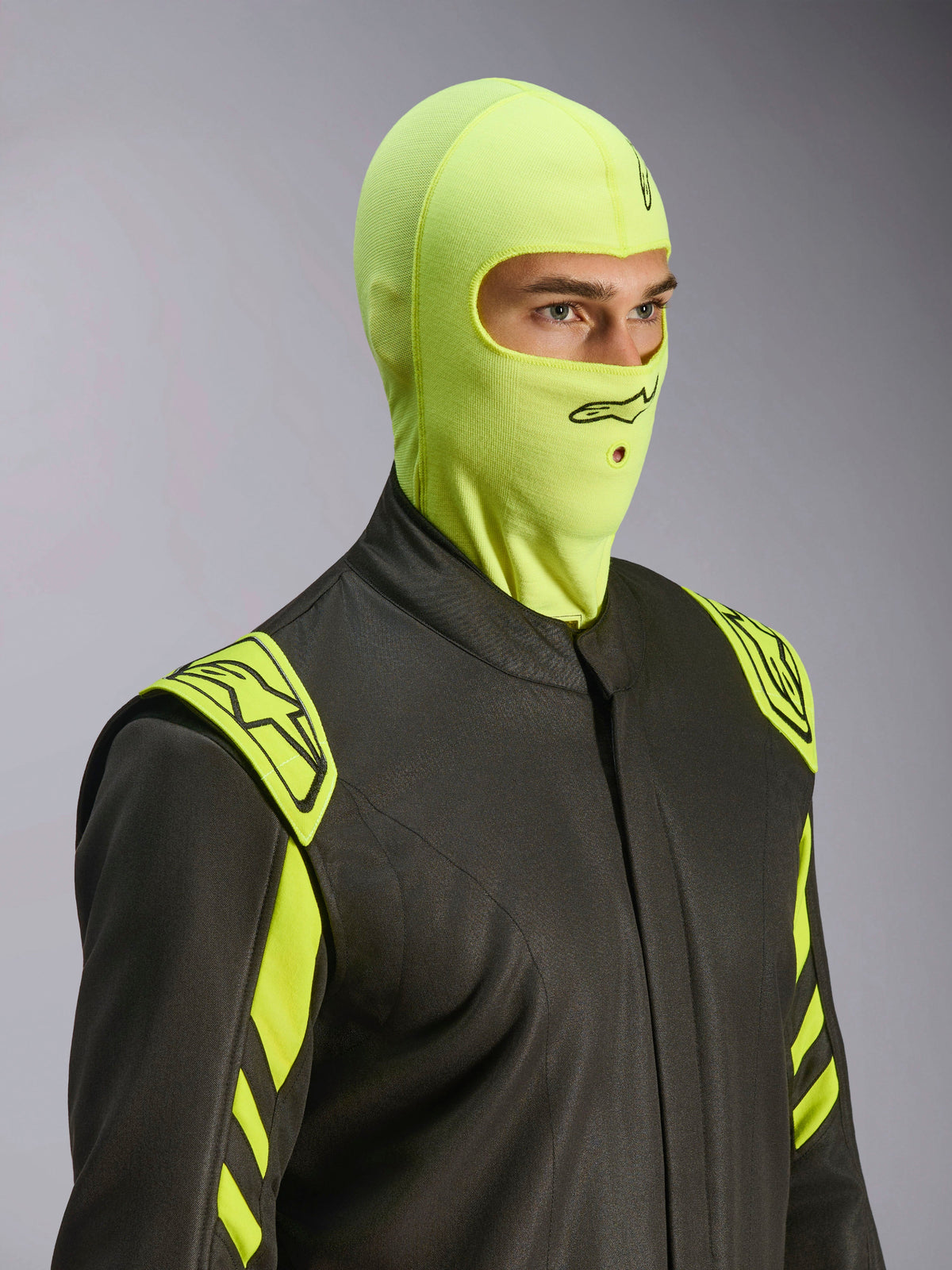 Male model wearing Alpinestars ZX Evo V3 Balaclava in Yellow Fluo, flame-resistant racing head sock, paired with a dark grey auto racing suit with neon yellow accents, featuring a black Alpinestars logo and mouth opening for hydration tube, professional motorsport protective gear.