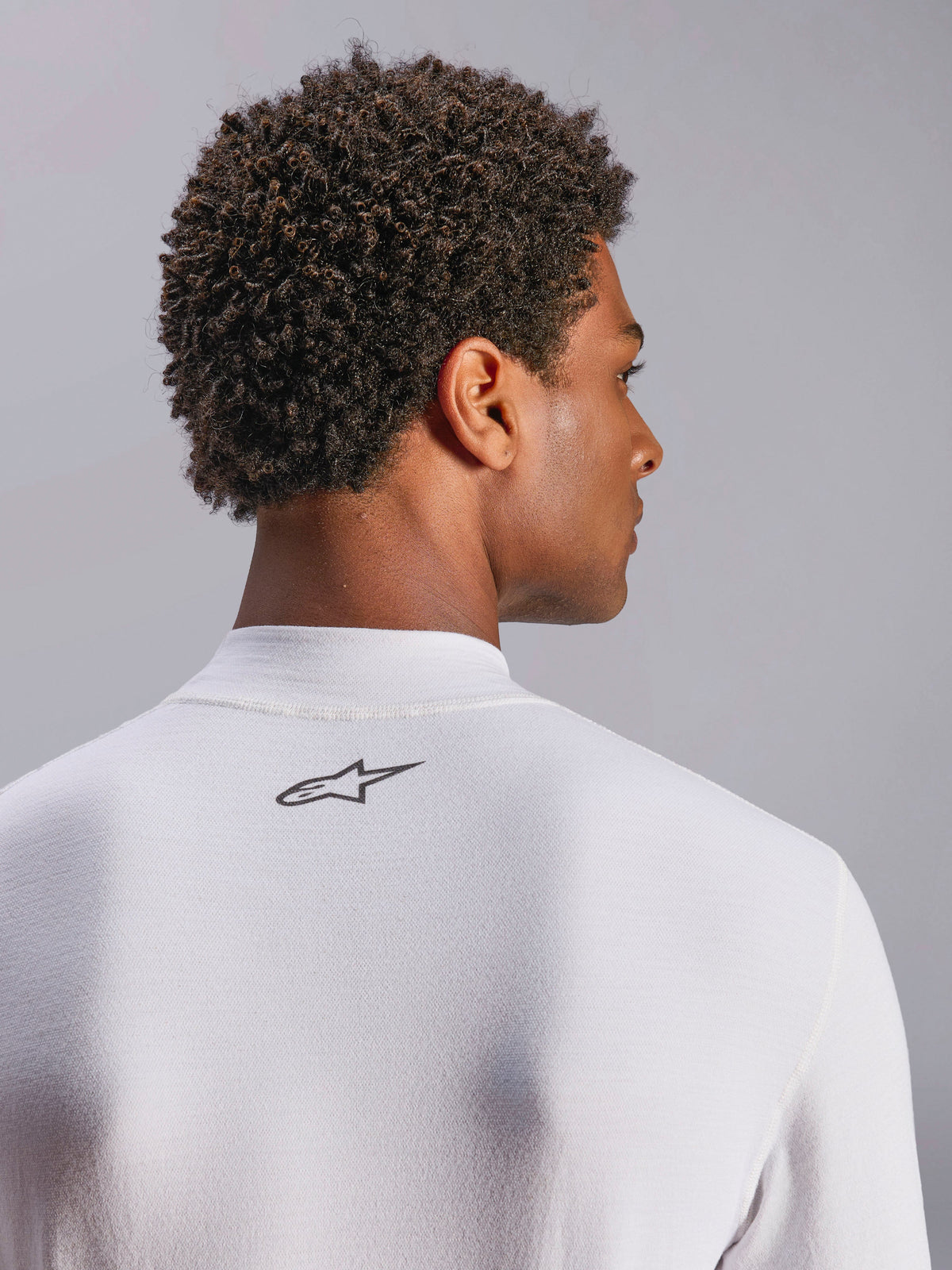 Alpinestars ZX Evo V3 Top, biała bielizna termoaktywna z długim rękawem do wyścigów samochodowych, zbliżenie na kark z czarnym logo Alpinestars, techniczna tkanina ognioodporna, model z ciemnymi kręconymi włosami