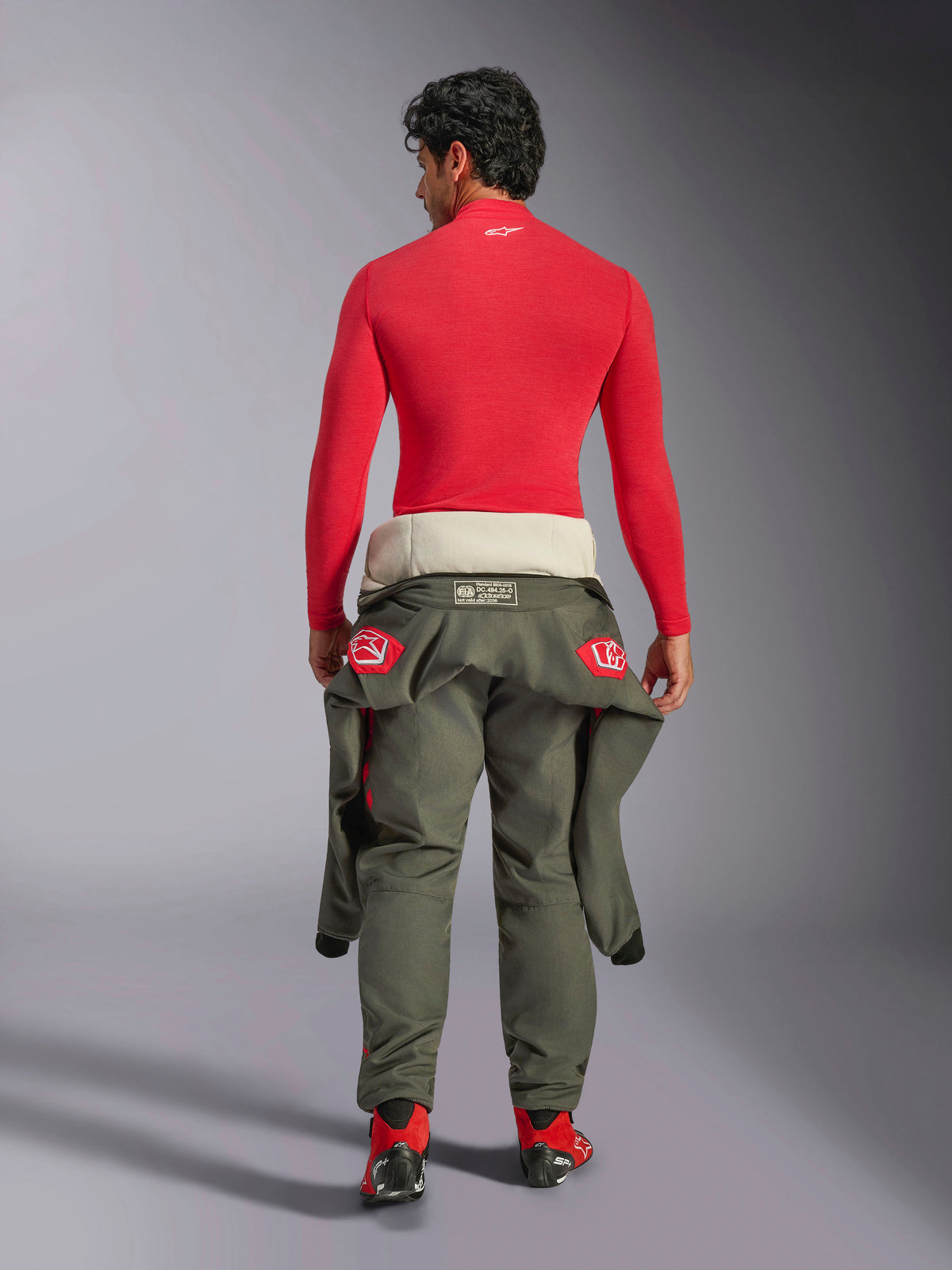 Alpinestars ZX Evo V3 Top - Long Sleeve, Red Auto Base Layer, back view of a male model wearing the fire-resistant top with olive green racing pants tied at the waist and red racing boots, professional motorsport gear.