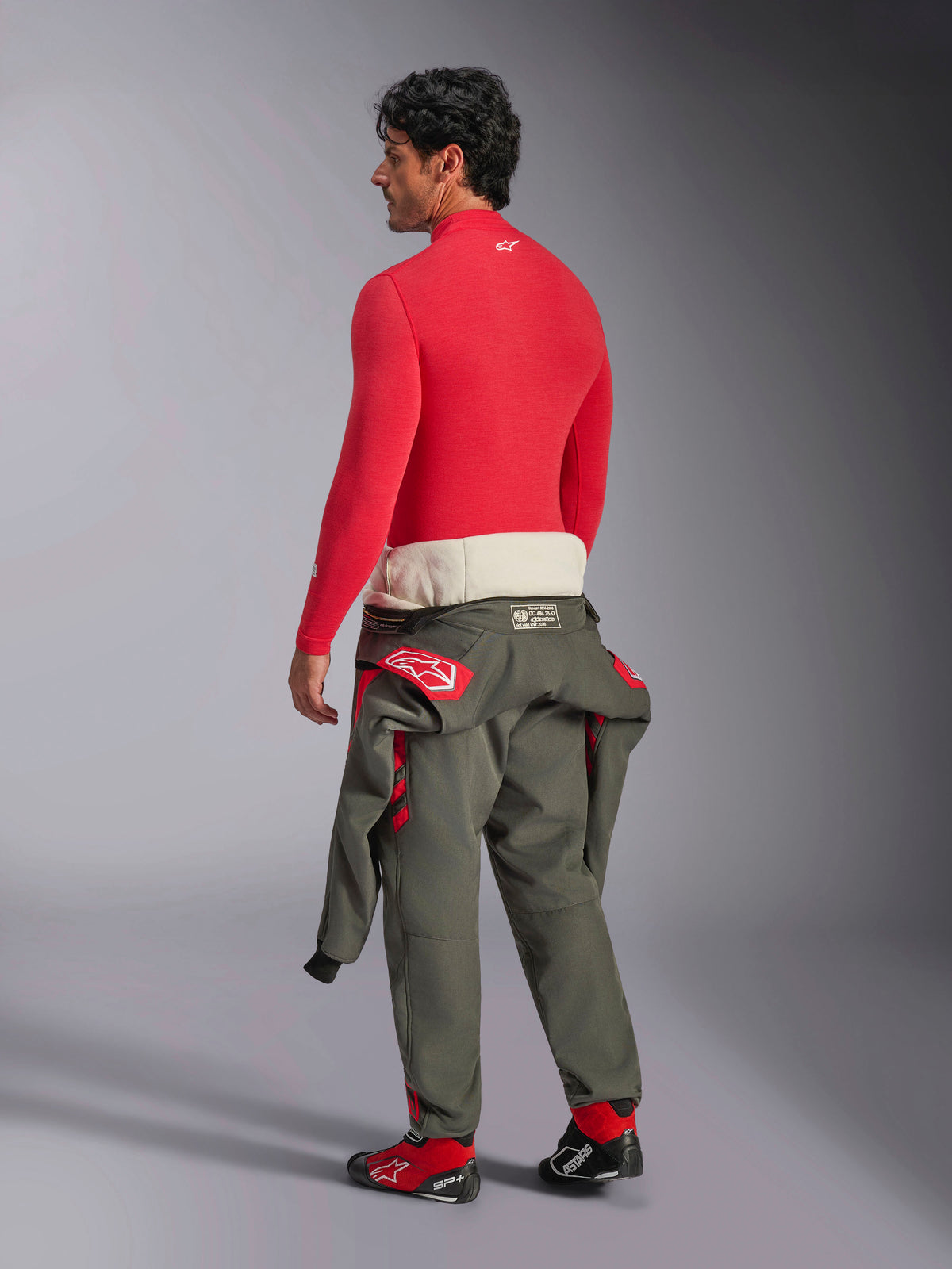 Alpinestars ZX Evo V3 Long Sleeve Top, Red auto base layer, back view on male model wearing olive green racing suit pants at the waist and red racing shoes, flame-resistant performance underwear for motorsport
