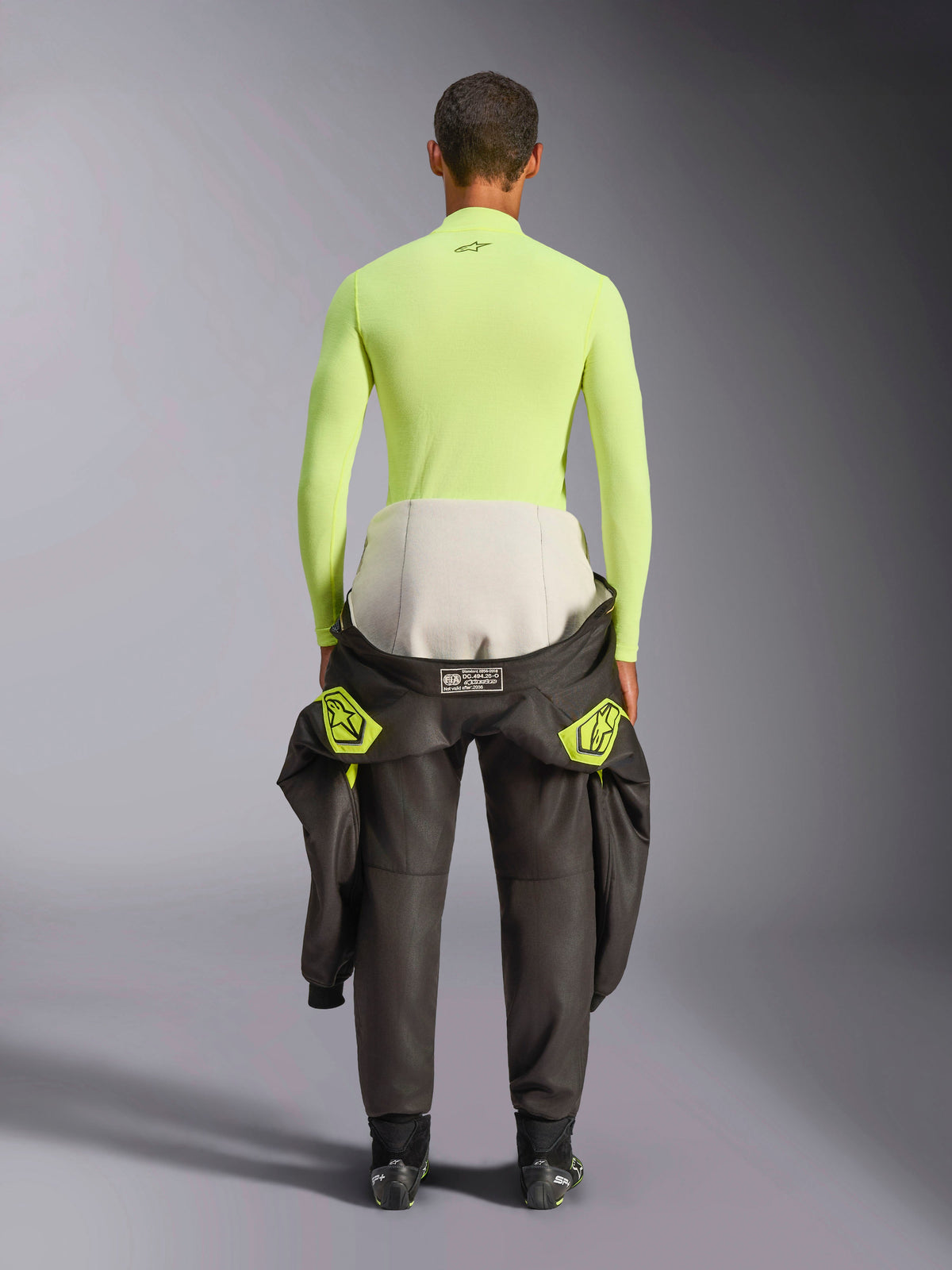 Alpinestars ZX Evo V3 Top, bielizna termoaktywna z długim rękawem, Yellow Fluo, widok od tyłu kierowcy z kombinezonem wyścigowym przewiązanym w pasie, jaskrawy neonowożółty ognioodporny top z małym czarnym logo na karku, dopasowany sportowy krój