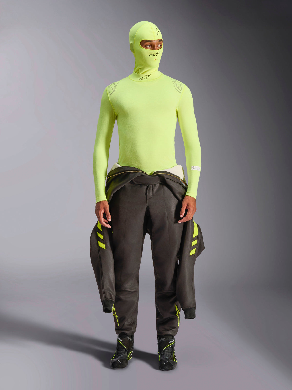 Alpinestars ZX Evo V3 Long Sleeve Top, żółty fluo, noszony z pasującą kominiarką i ciemnym kombinezonem wyścigowym przewiązanym w pasie, lekka ognioodporna warstwa bazowa do wyścigów samochodowych, z logo Alpinestars na kołnierzu i ramionach