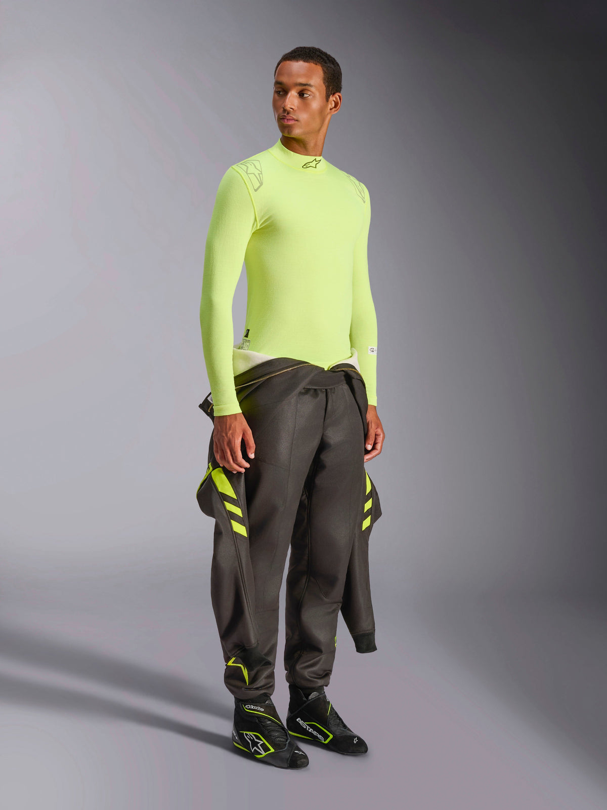 Alpinestars ZX Evo V3 Top - Long Sleeve, żółty fluo, noszony przez modela z ciemnoszarym kombinezonem wyścigowym przewiązanym w pasie i czarnymi butami wyścigowymi, ognioodporna bielizna termoaktywna do sportów motorowych, lekka konstrukcja z siatki żakardowej, profesjonalny sprzęt do wyścigów samochodowych