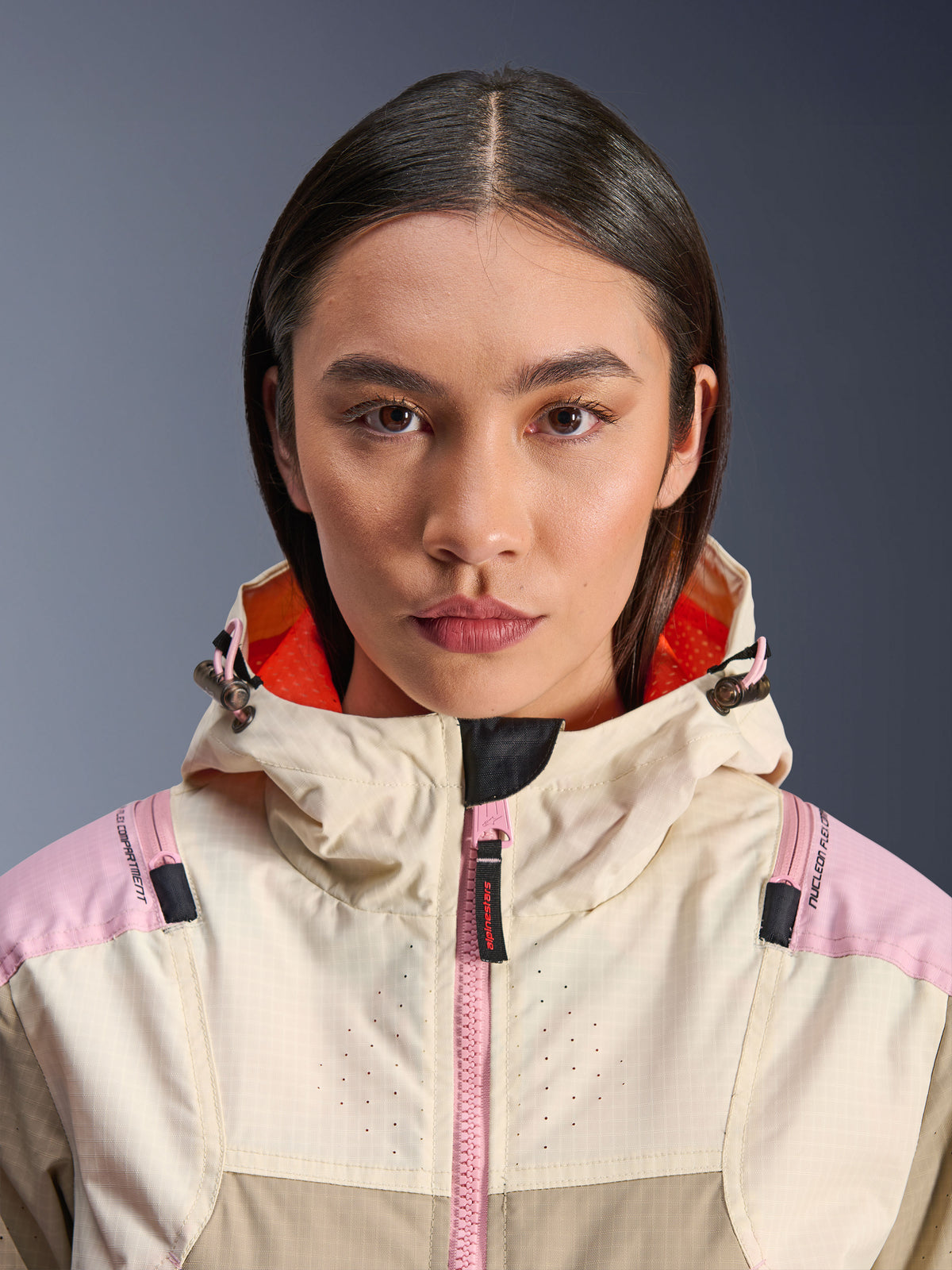Zbliżenie na modelkę w kurtce Alpinestars Molly Air Jacket w kolorze Sand Pink Almond, z wysokim kołnierzem i kapturem z jaskrawopomarańczową podszewką, różowy zamek, materiał ripstop z laserową perforacją na klatce piersiowej dla wentylacji, miejska motocyklowa odzież ochronna