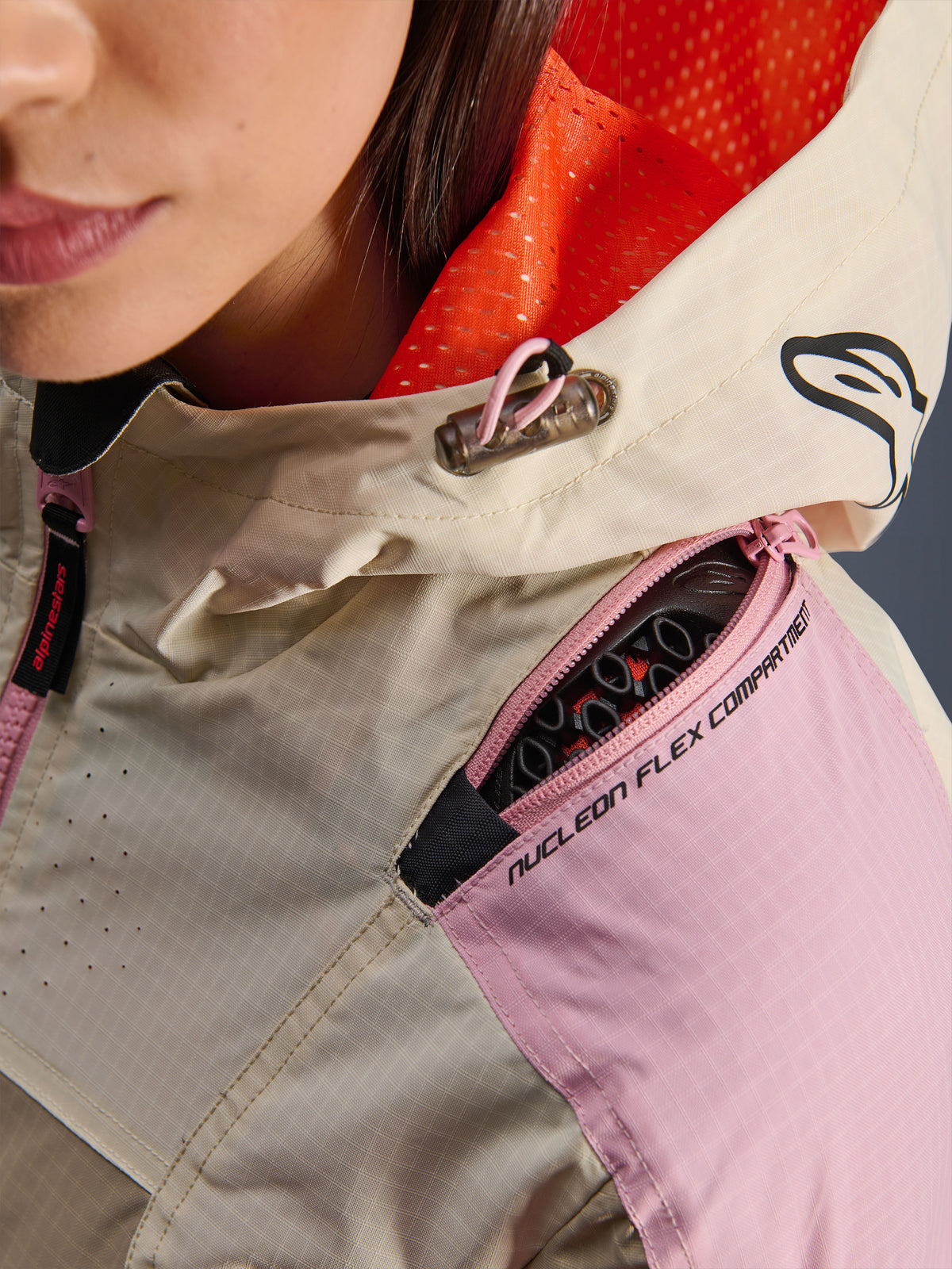 Zbliżenie na kurtkę Alpinestars Molly Air w kolorze Sand Pink Almond, prezentujące zapinaną na zamek kieszeń na ramieniu na ochraniacze Nucleon Flex, jaskrawopomarańczową podszewkę kaptura z siateczki oraz wytrzymałą konstrukcję z materiału ripstop.