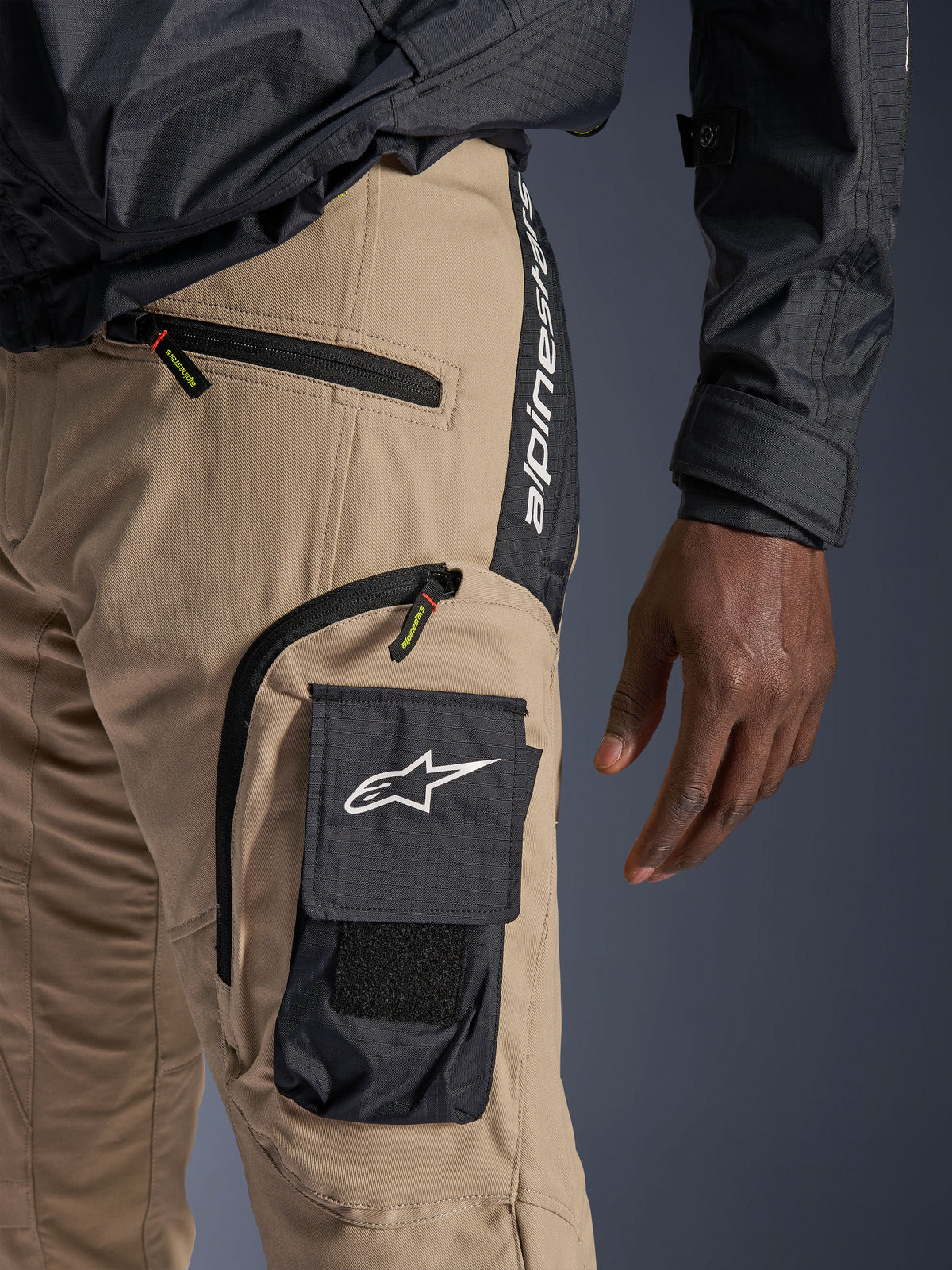 Spodnie Alpinestars Acteon w kolorze Sand, szczegółowy widok okolicy bioder i uda, czarna boczna kieszeń cargo z białym logo gwiazdy, kieszenie zapinane na zamek, pionowy napis Alpinestars na czarnym panelu bocznym, miejska odzież motocyklowa