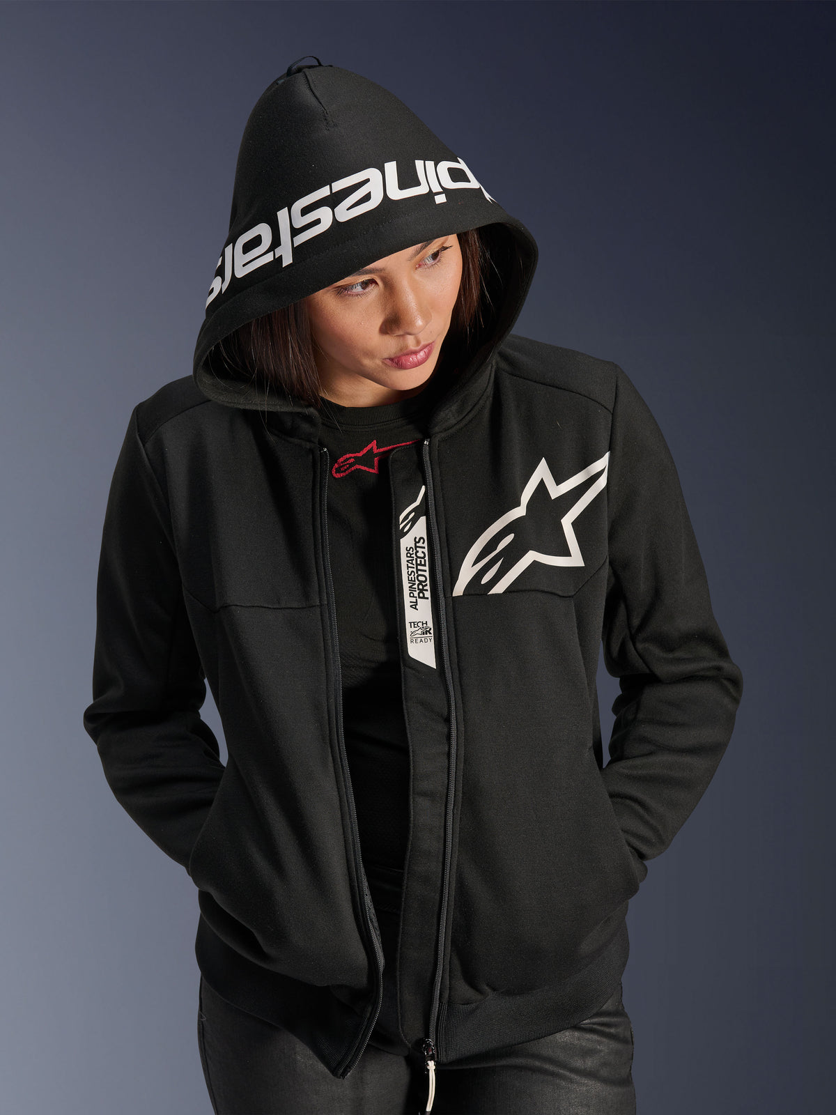 Alpinestars Stella Chrome V2 Sport Hoodie, damska bluza motocyklowa, czarno-biała, białe logo gwiazdy na piersi, branding Alpinestars na krawędzi kaptura, kobieta z założonym kapturem, swobodny miejski styl sportowy, detale Tech-Air Ready