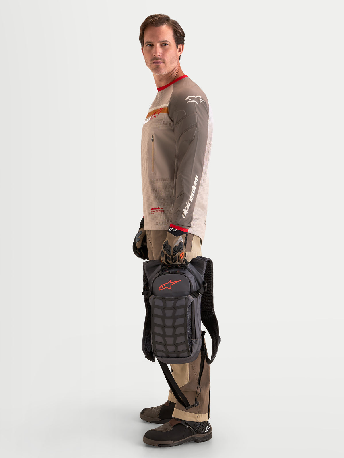 Alpinestars Plecak AT-9, plecak motocyklowy motocross w kolorze brązowym i ciemnoszarym, widok z boku na modela stojącego i trzymającego lekki plecak za górny uchwyt, prezentujący panel strukturalny i zorganizowane przegrody