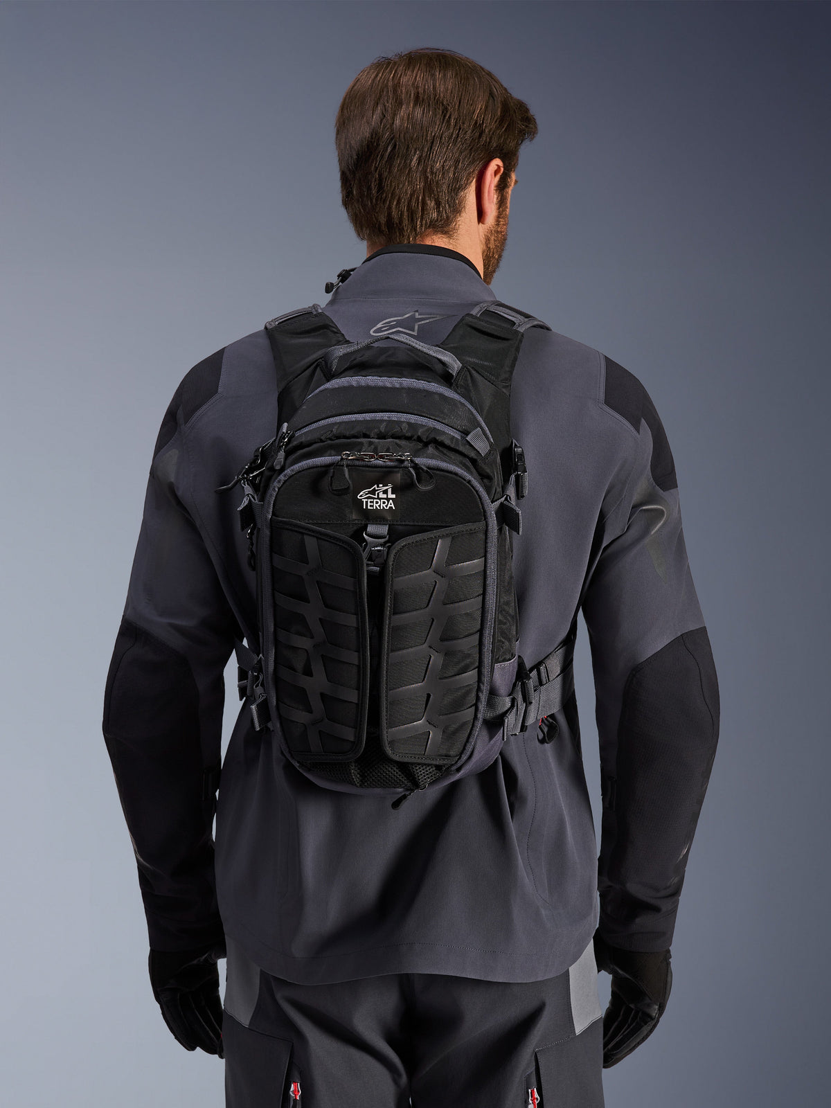 Alpinestars AT-15 Back Pack, przygodowy plecak motocyklowy, Black Deep Gray, noszony przez osobę na szarej technicznej kurtce motocyklowej, widok z tyłu z brandingiem „ALL TERRA”, ergonomiczny design ze wzmocnionymi teksturowanymi panelami i solidnymi pasami.