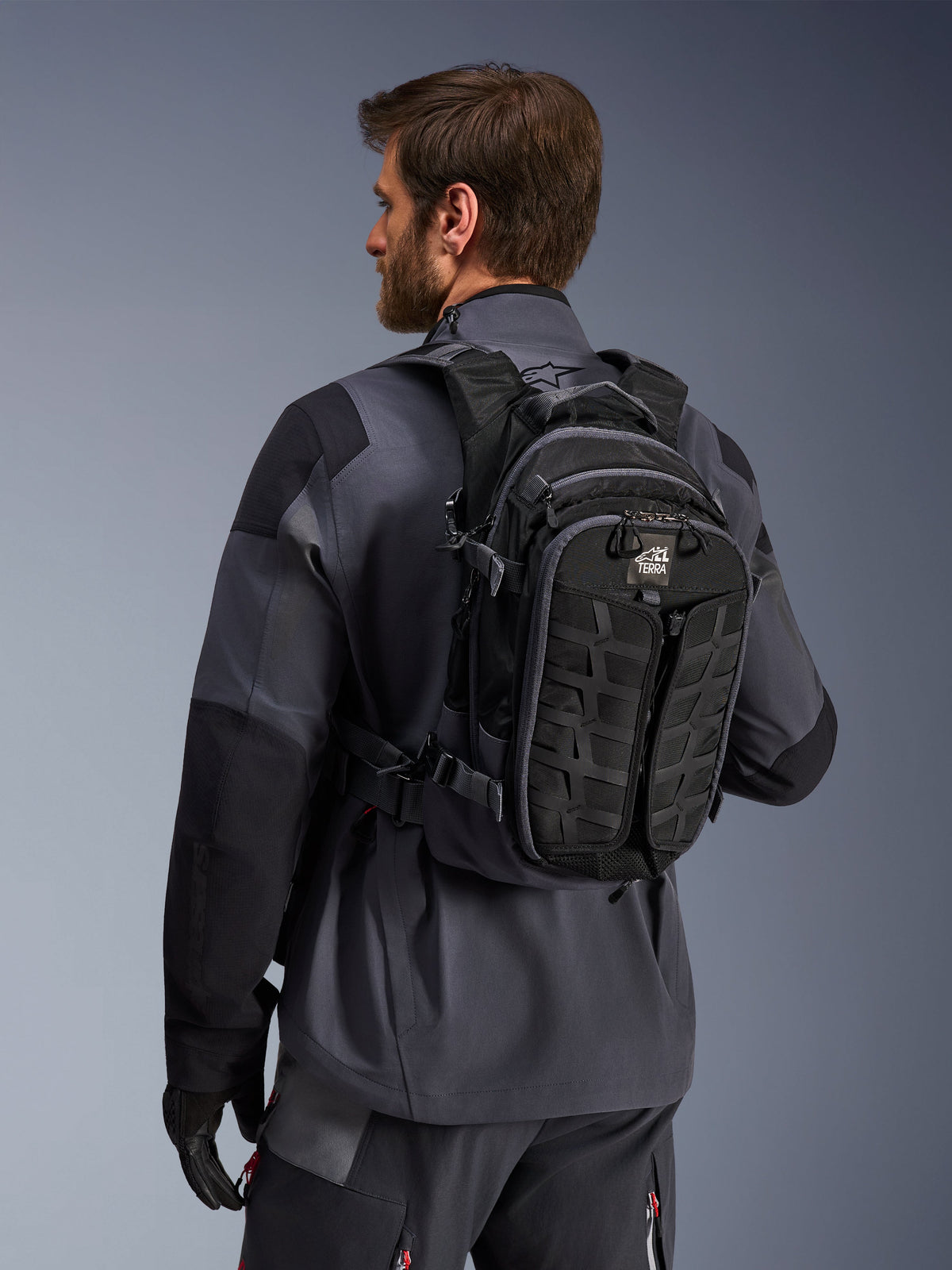 Alpinestars AT-15 Back Pack, motocyklowy plecak adventure, czarny i ciemnoszary, noszony przez motocyklistę w szarej kurtce, o technicznym designie z systemem MOLLE i logo „TERRA”, ergonomiczny sprzęt off-road