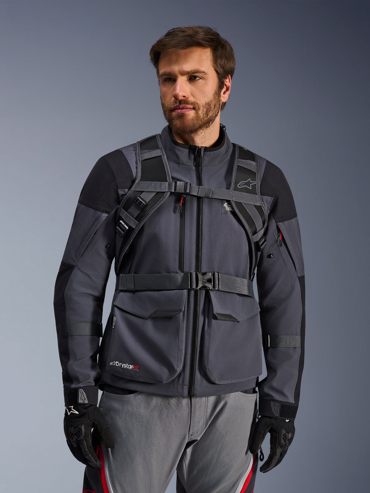 System szelek plecaka Alpinestars AT-15, Black Deep Gray, widok z przodu na motocykliście w szarej kurtce motocyklowej, wyposażony w magnetyczne zapięcie na klatce piersiowej i pas biodrowy, czarne rękawice, styl adventure