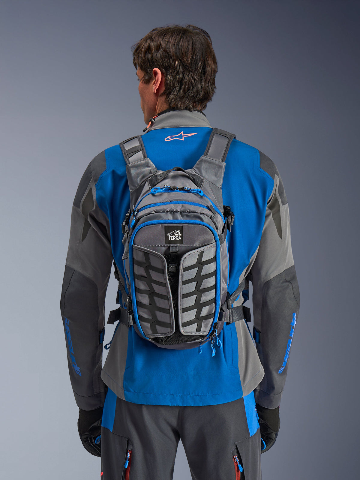 Alpinestars AT-15 Back Pack, adventure motorcycle backpack, Dark Gray and Deep Gray, worn by a rider over a blue and gray jacket, featuring All Terra branding, ergonomic structural design, adjustable straps, and blue zipper accents.