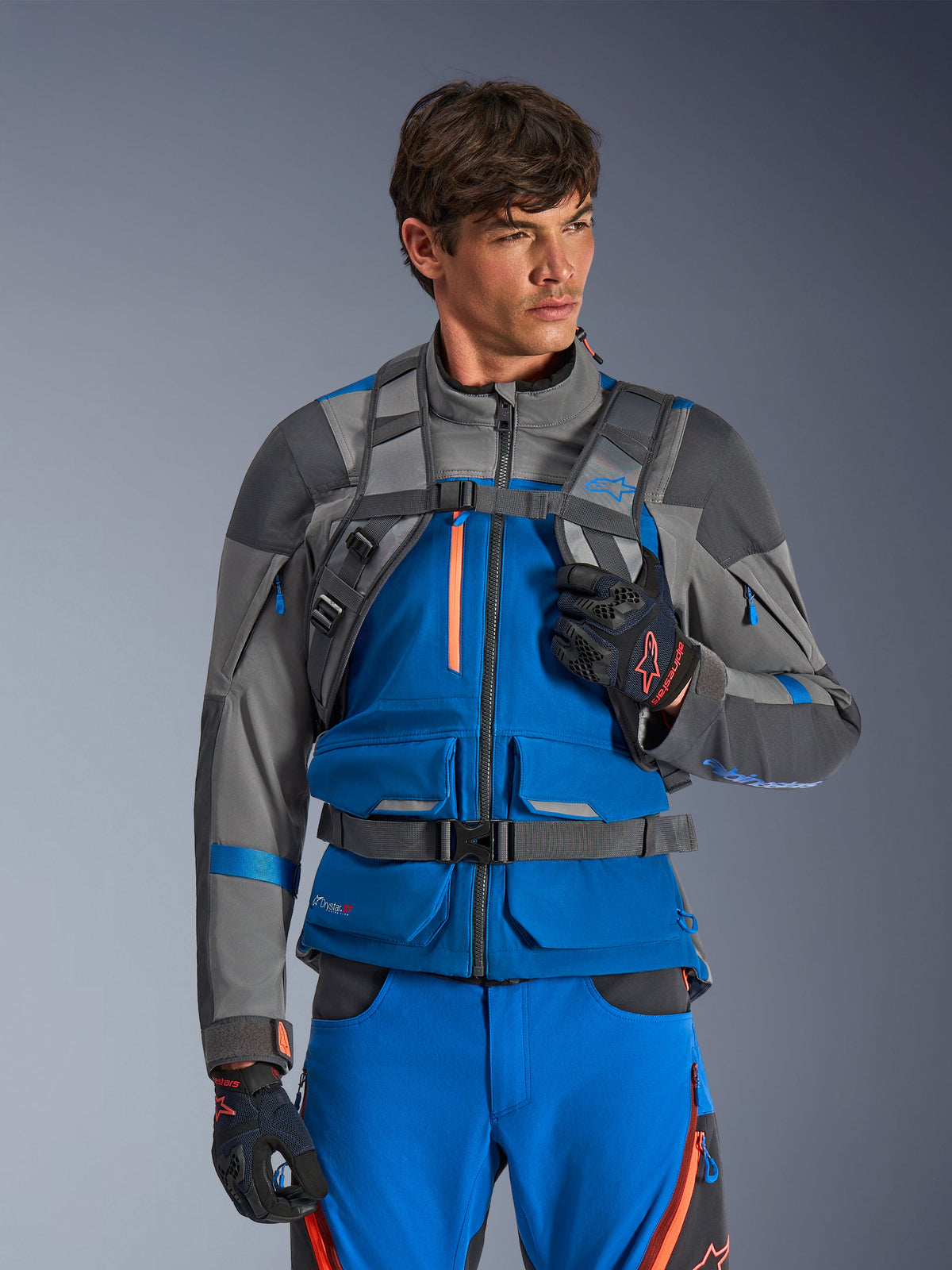 Alpinestars AT-15 Back Pack, Motorcycle Backpack, Dark Gray, worn by a rider in a blue and gray adventure jacket and pants, featuring technical gray shoulder straps with magnetic chest buckle and waist strap, rugged design for off-road riding