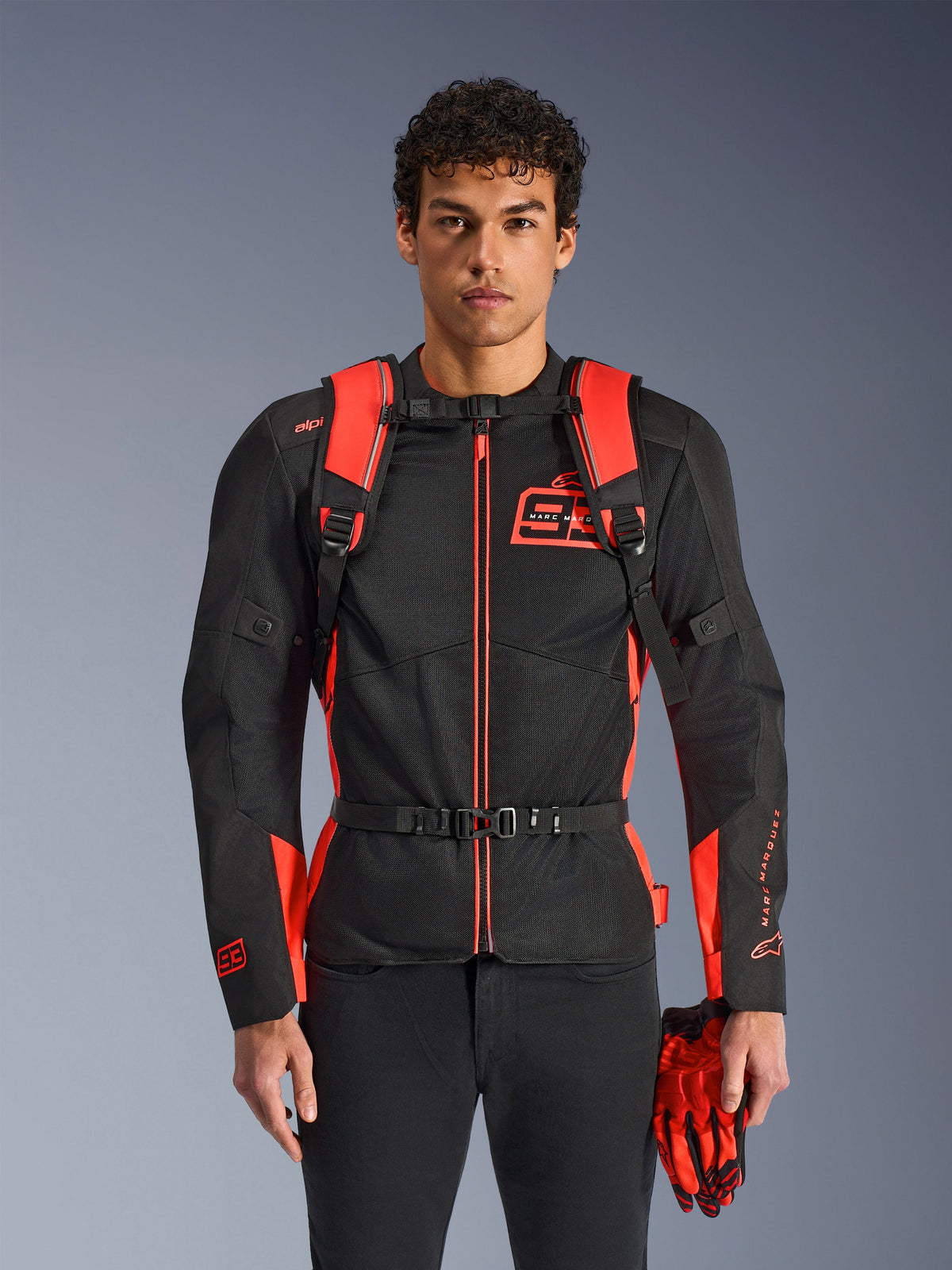Man wearing Alpinestars MM93 AMP3 Backpack, Red Fluo and Black, featuring adjustable shoulder straps and buckled chest strap, styled with a matching MM93 motorcycle jacket and gloves, technical gear from the Marc Marquez collection