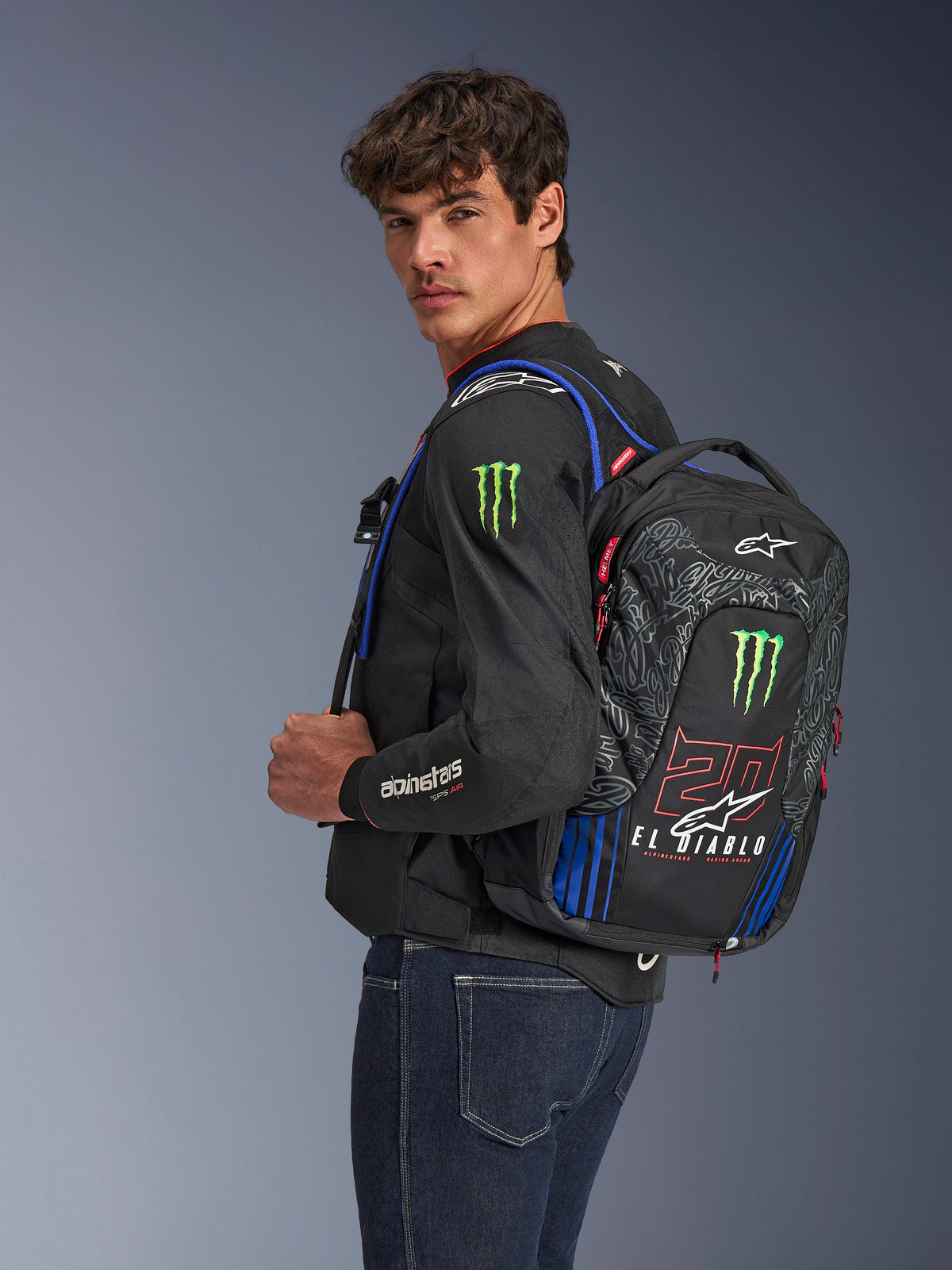 Alpinestars FQ20 City Hunter V2 Monster Backpack, plecak motocyklowy, czarny z akcentami w kolorze czerwonym fluo, niebieskim i zielonym, z logo Monster Energy oraz brandingiem Fabio Quartararo „El Diablo”, prezentowany na modelu w czarnej kurtce motocyklowej i jeansach, opływowy design dla motocyklistów miejskich