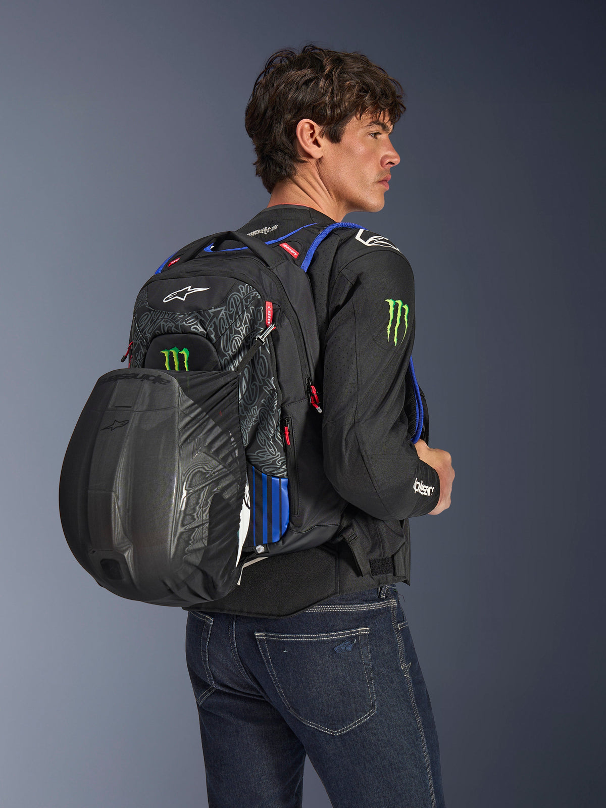Alpinestars FQ20 City Hunter V2 Monster Backpack, miejski plecak motocyklowy, czarny z czerwonymi fluo, niebieskimi i zielonymi akcentami, model z czarnym kaskiem zamocowanym w siatce, czarna kurtka motocyklowa i niebieskie jeansy.