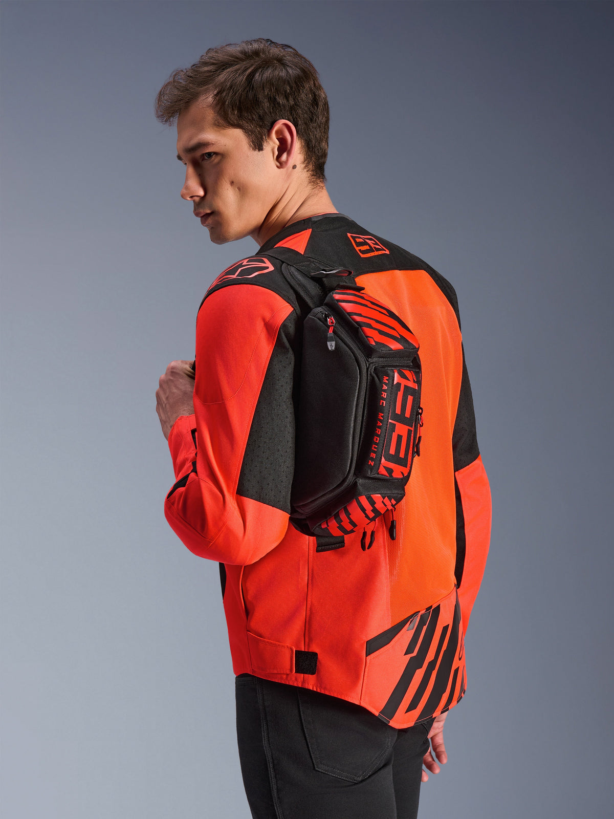 Torba na pas Alpinestars MM93, czarna z jaskrawoczerwonymi (Red Fluo) i jasnoczerwonymi (Bright Red) elementami, noszona przez modela na plecach na czerwono-czarnej kurtce motocyklowej, z wyraźnym brandingiem MM93 i grafikami Marca Marqueza