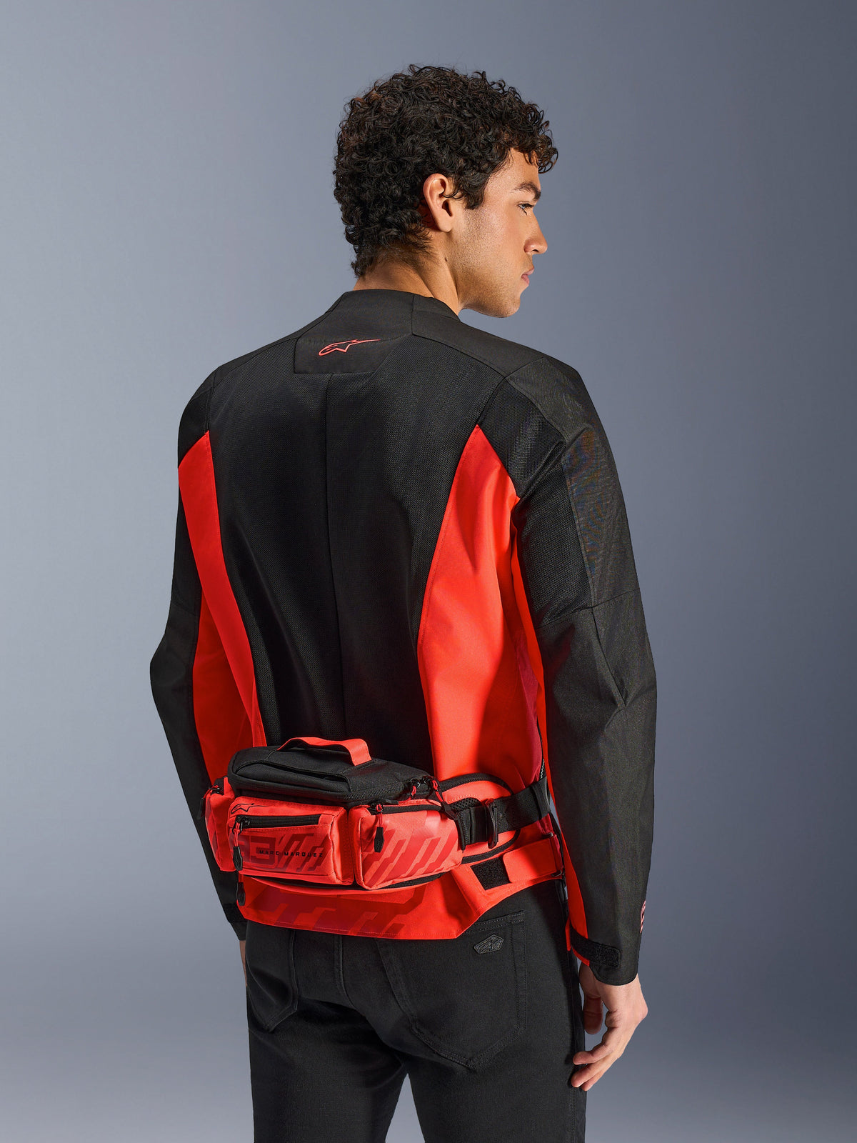 Alpinestars MM93 Waist Bag, motocyklowa torba na pas, jaskrawoczerwona i czarna, noszona na dopasowanej czarno-czerwonej kurtce motocyklowej, posiada przegrody na suwak oraz branding Marc Marquez MM93, wytrzymała konstrukcja tekstylna do jazdy na motocyklu