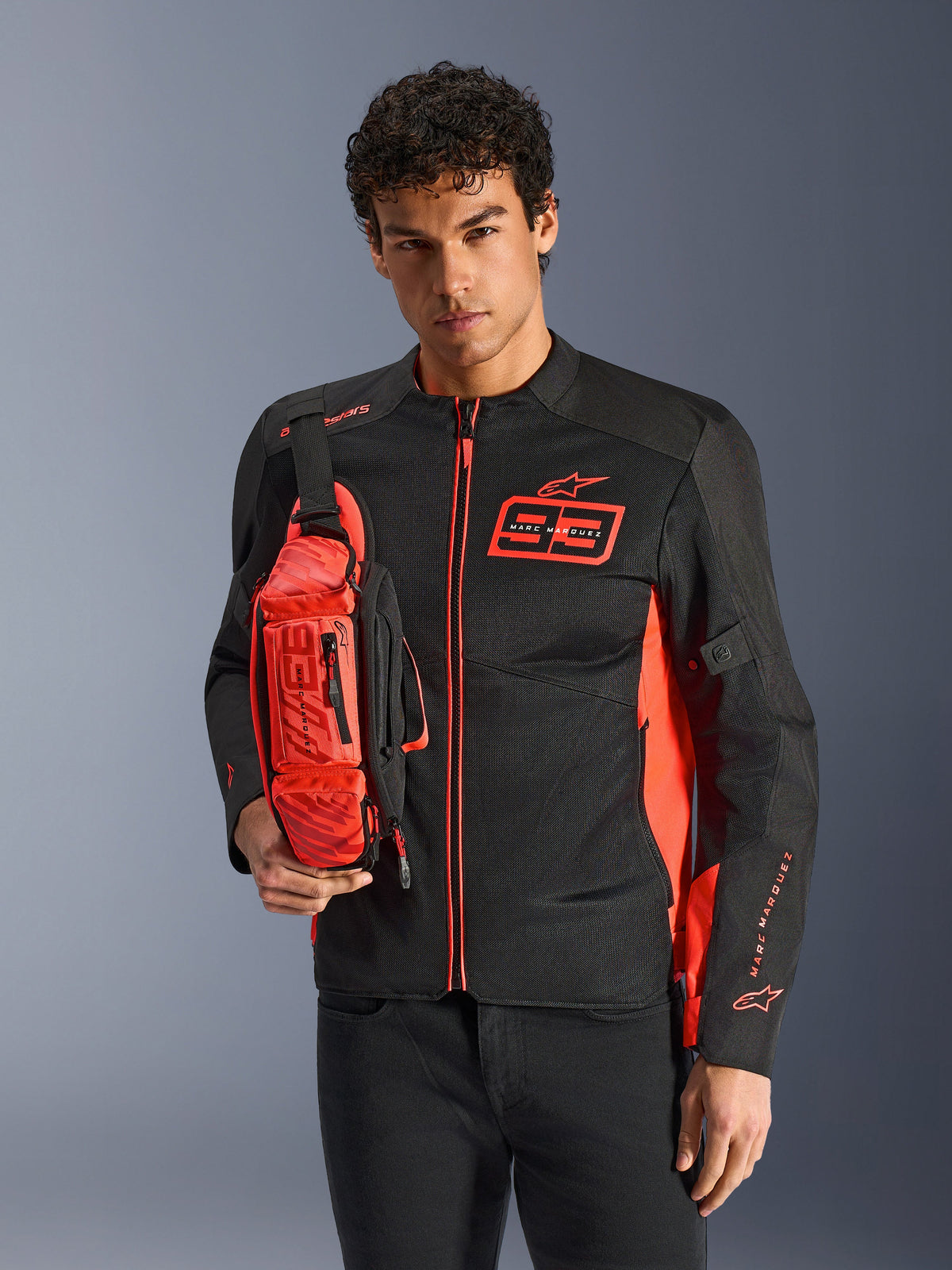 Mężczyzna noszący torbę na pas Alpinestars MM93, w kolorze Red Fluo i czarnym, stylizowaną na torbę na ramię na dopasowanej czarno-czerwonej kurtce motocyklowej, z logo Marc Marquez 93, sportowy design wyścigowy