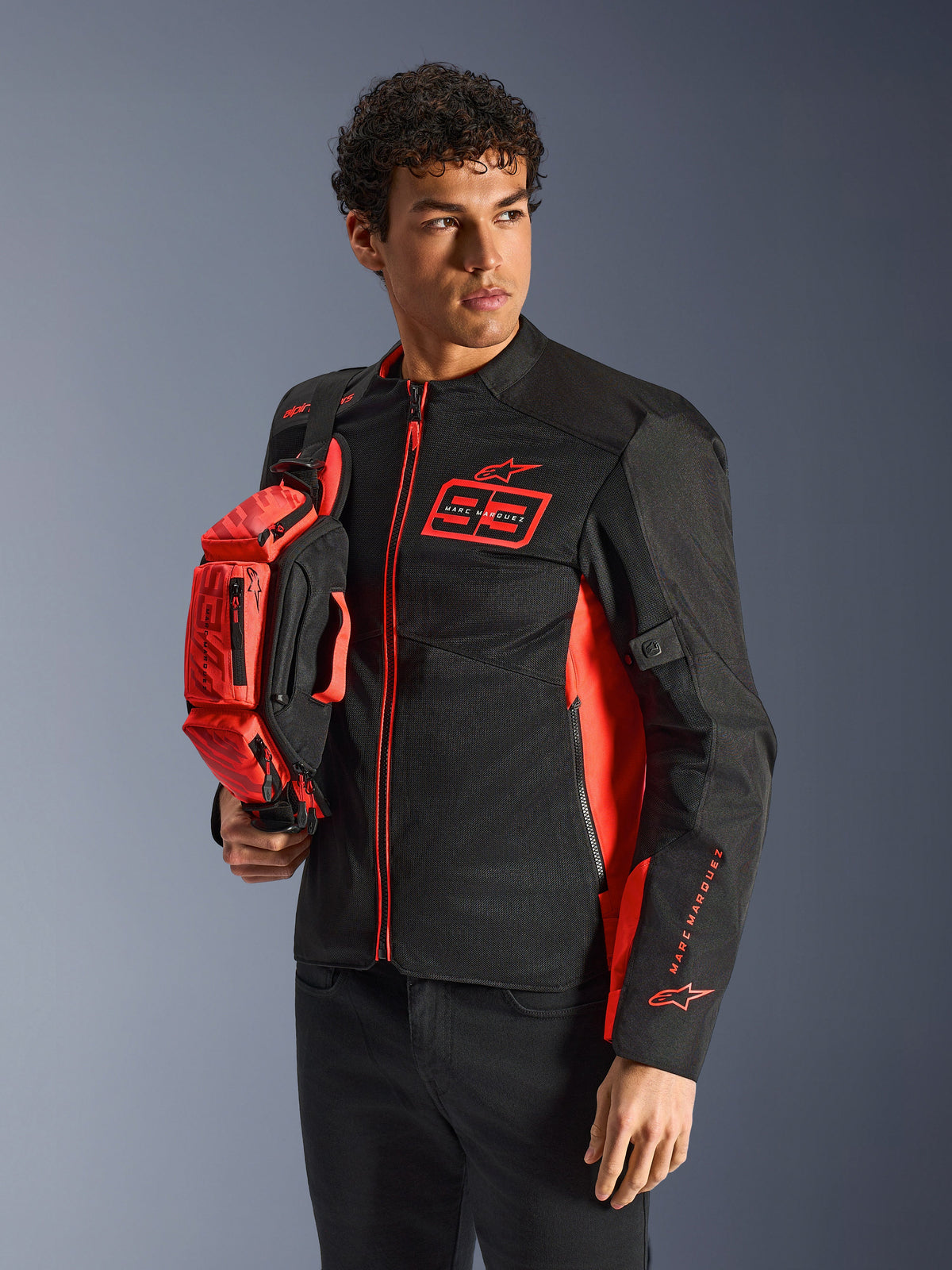 Alpinestars MM93 Waist Bag, torba na pas w kolorze czerwonym fluo i czarnym, trzymana przez modela w pasującej czarno-czerwonej kurtce motocyklowej, wyposażona w wiele przegród na zamek i branding Marca Marqueza, praktyczne wyposażenie do jazdy sportowej