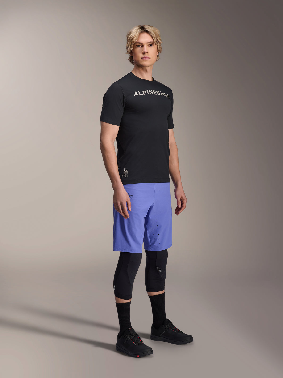 Męski model w odzieży rowerowej MTB Alpinestars, czarna koszulka A-Aria Switch SS i niebieskie szorty A-Aria Lite Baja Blue, czarne ochraniacze kolan, czarne skarpetki i czarne buty rowerowe MTB, ujęcie studyjne całej sylwetki.