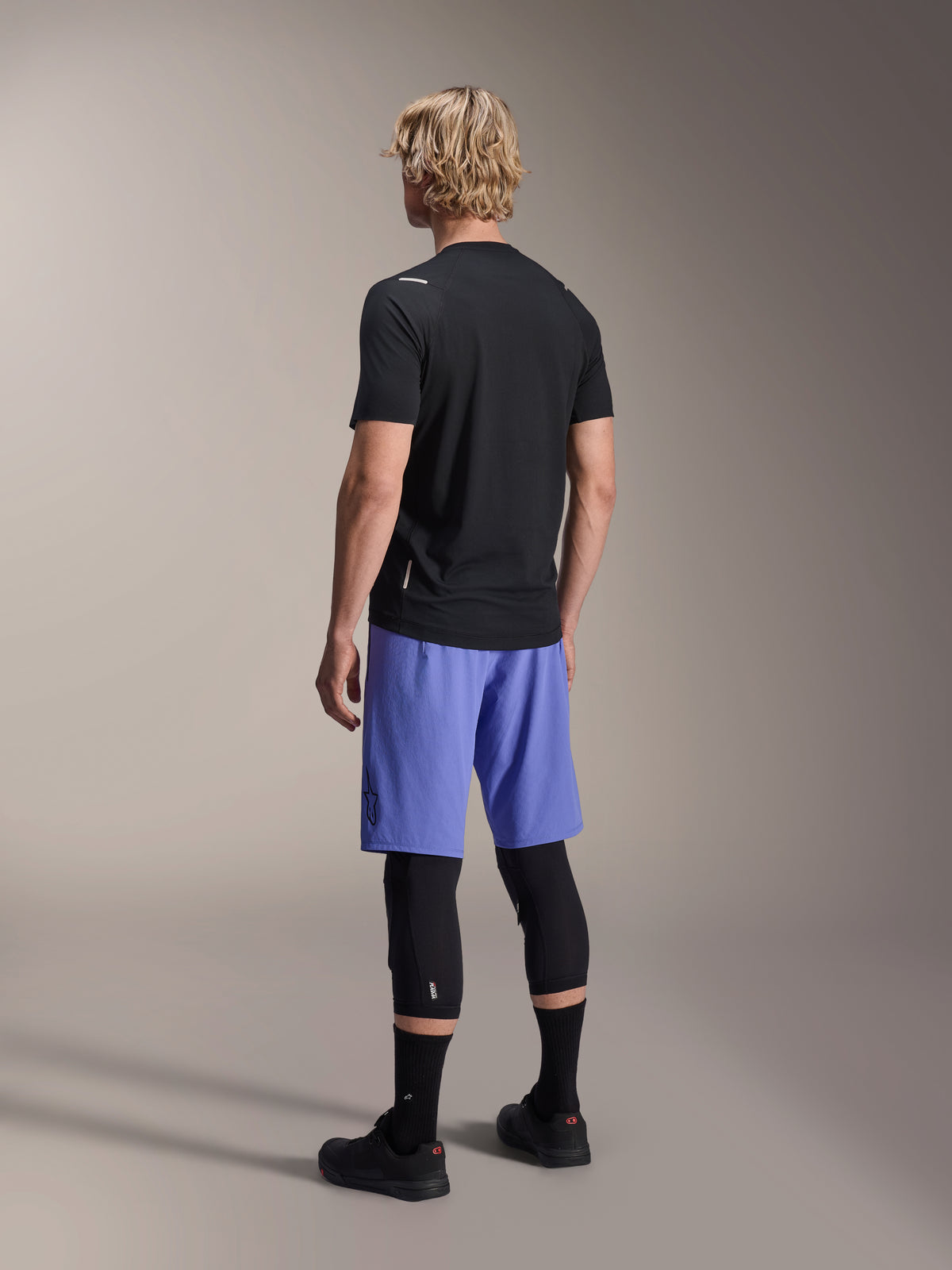 Model od tyłu w spodenkach Alpinestars A-Aria Lite w kolorze Baja Blue, zestawionych z czarną koszulką z krótkim rękawem i czarnymi ochraniaczami kolan, profesjonalna odzież rowerowa MTB do jazdy po szlakach.